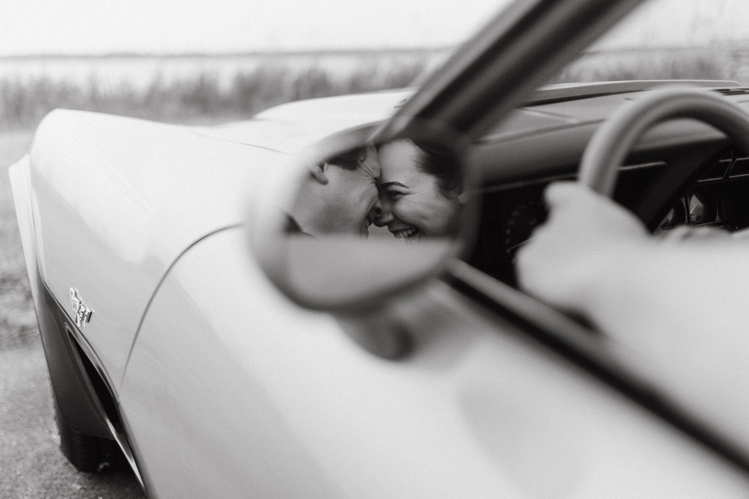 Black and white photo of a couple sharing a romantic kiss inside a vintage convertible car.