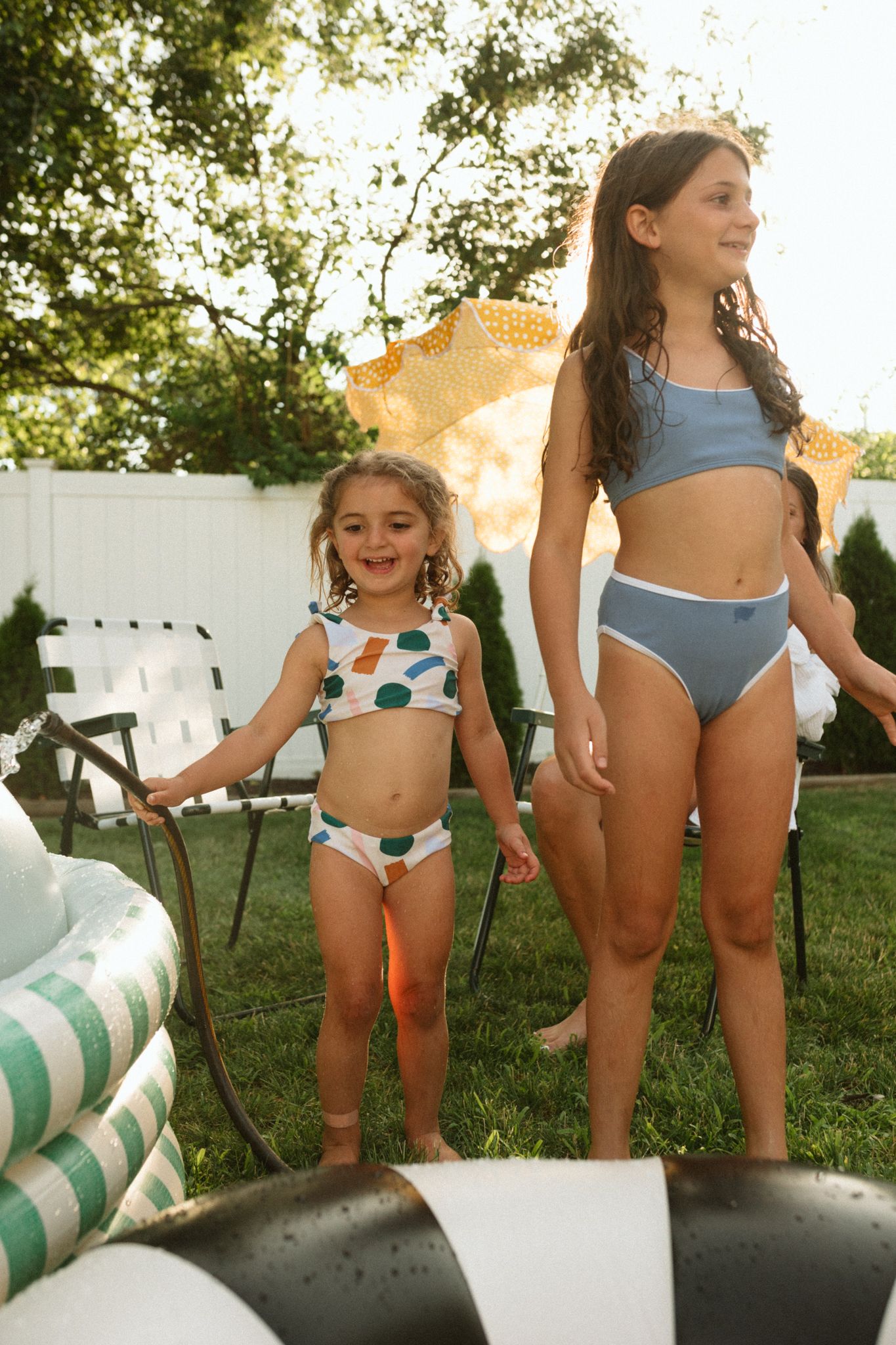 Two figures standing on pool floats in a sunny backyard with white fence.