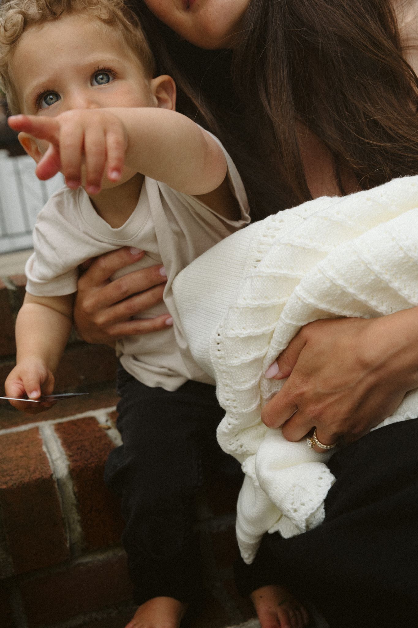 A mother holds a curious toddler with blue eyes on brick steps, as baby reaches out pointing at something nearby.