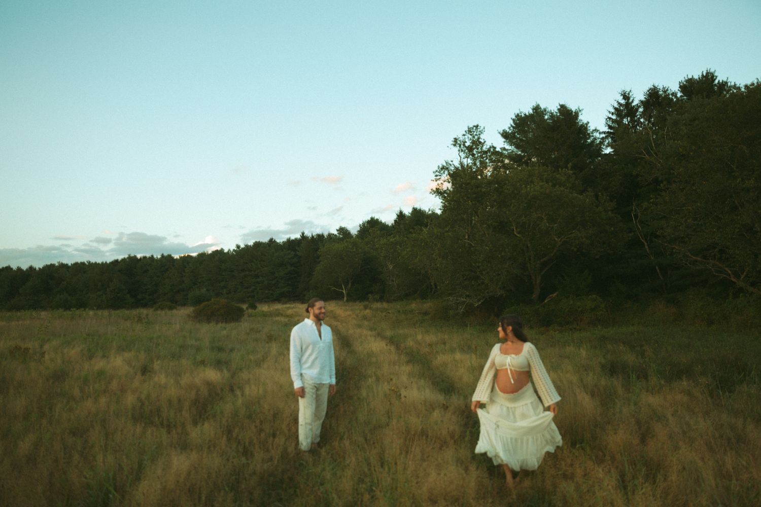 Couple in white attire walking through a sunlit meadow with lush green trees in the background at dusk.