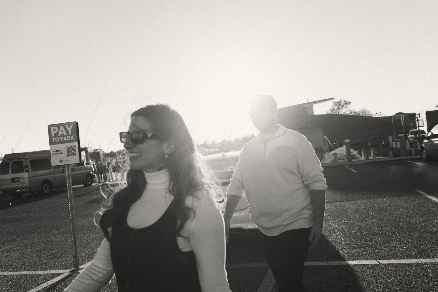Black and white photo of a couple walking in a sunny parking lot, woman in sunglasses foreground, man behind her.