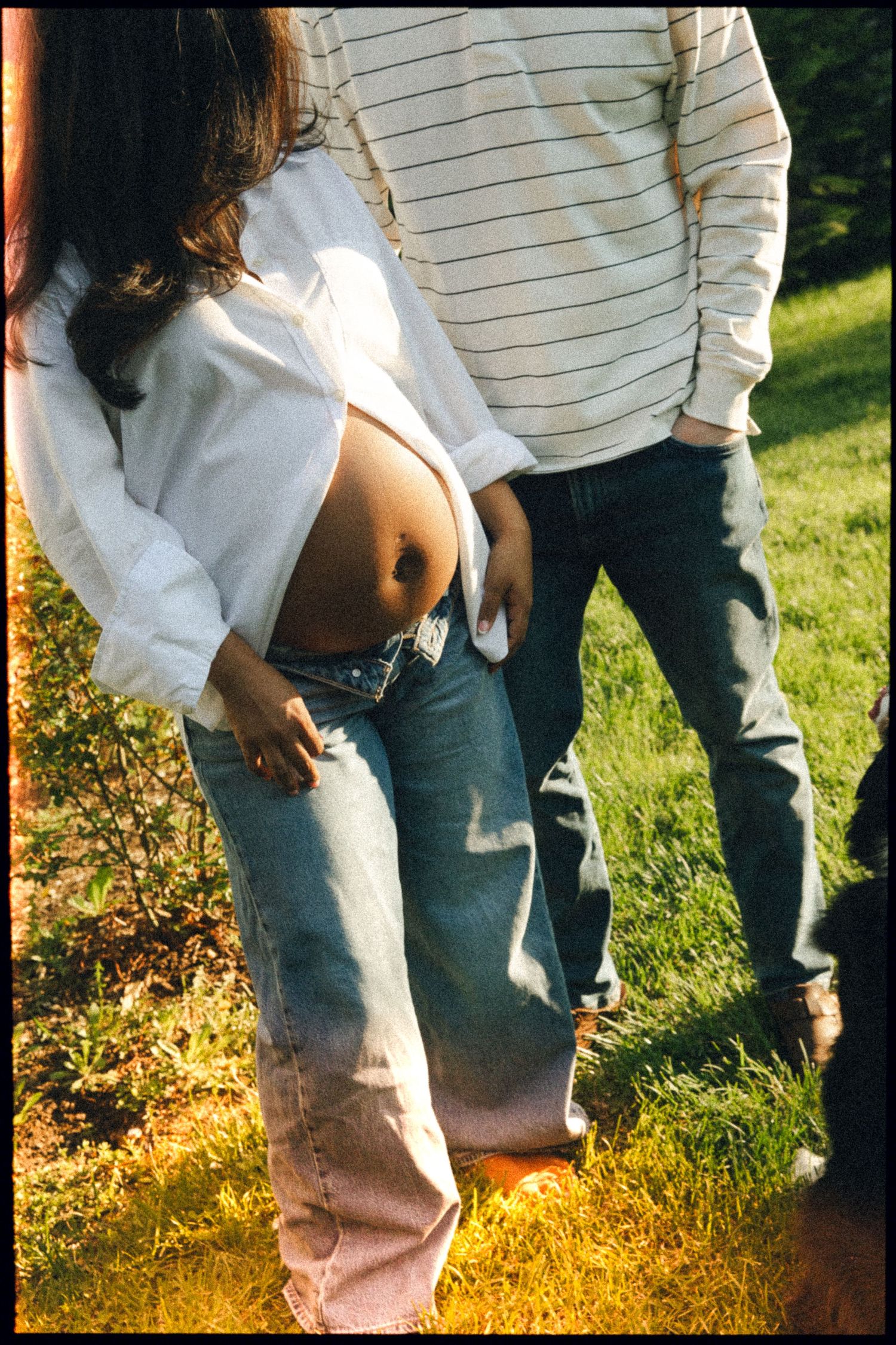 Pregnant woman in white shirt and jeans stands outdoors with partner, showing baby bump in sunny garden setting.