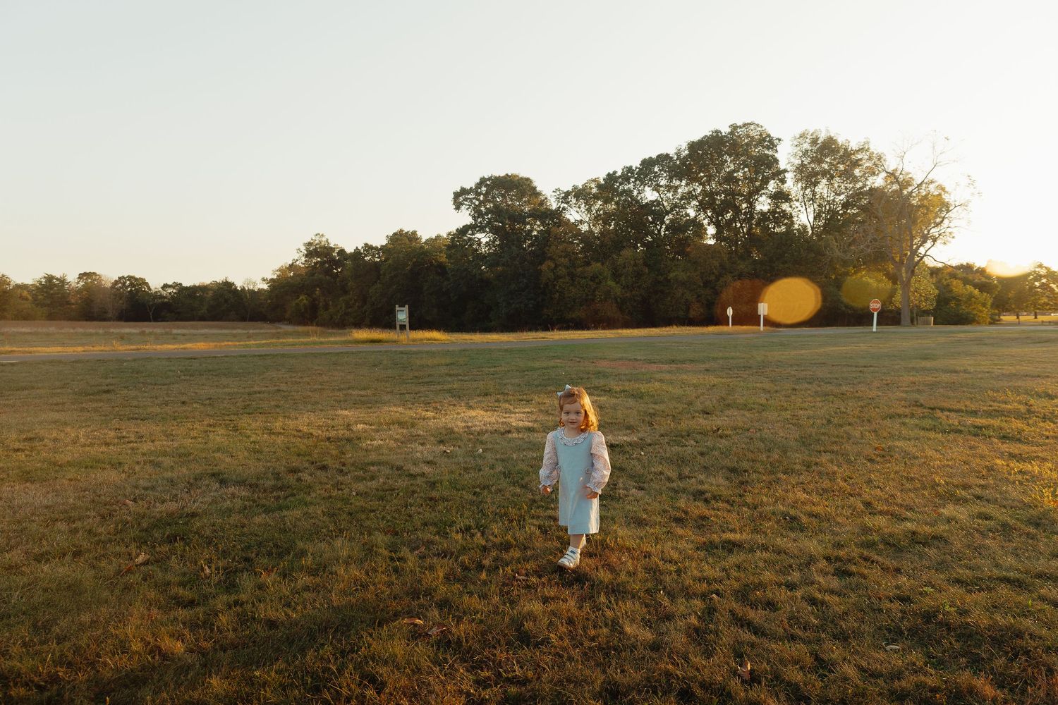 A young child in a white outfit runs through a sunlit green field at golden hour with trees in the background.