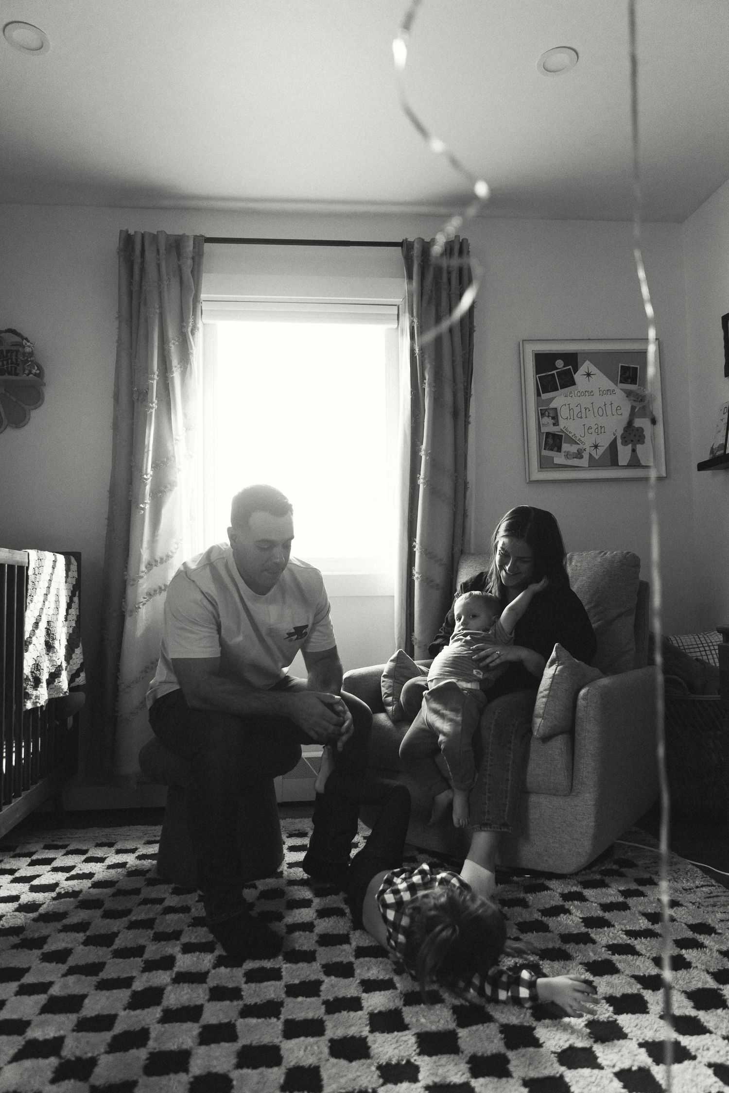 Black and white photo of a family with a baby relaxing in a cozy living room with string lights.