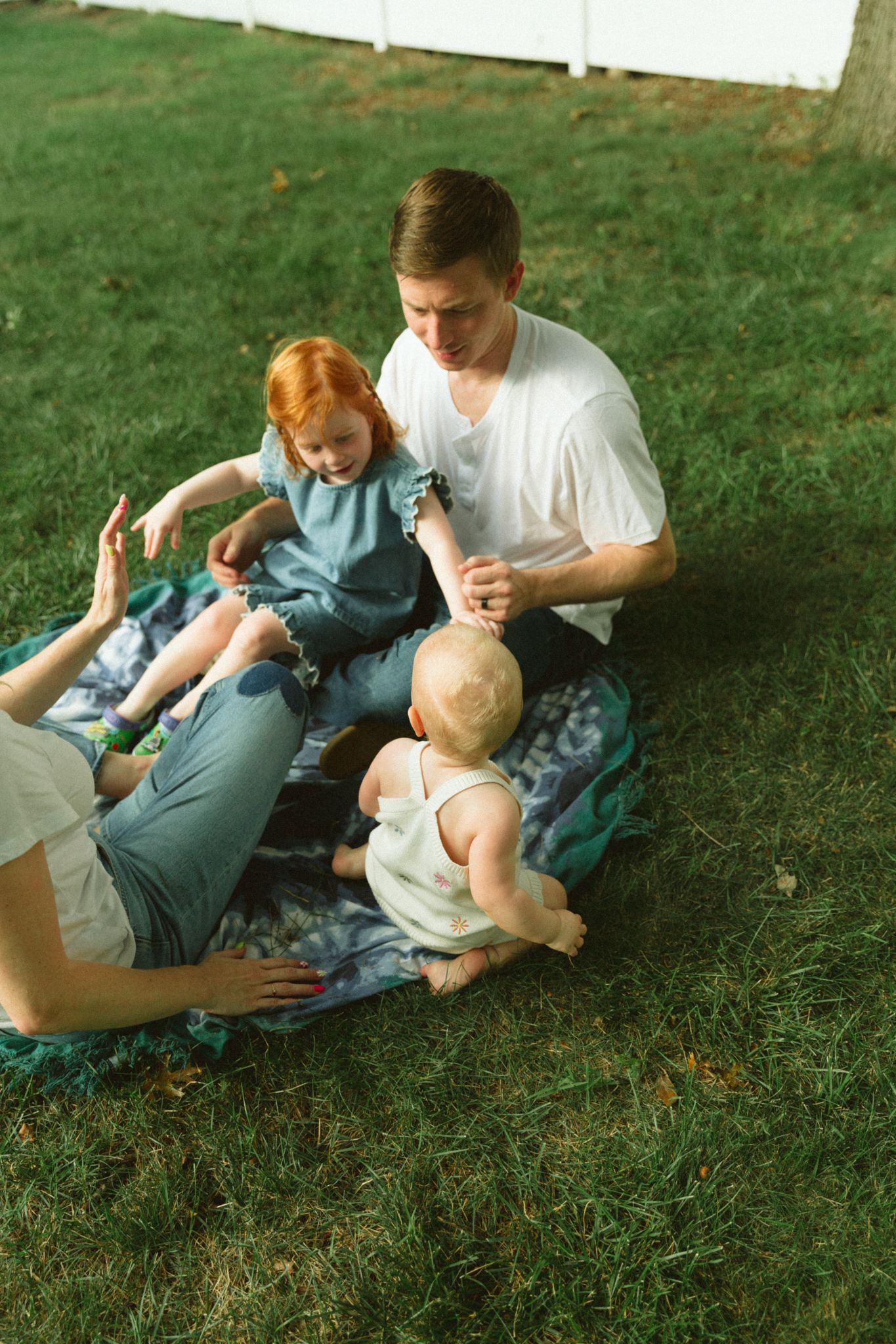 A young father sits on a blanket in green grass playing with two small children, a toddler girl and baby, outdoors.