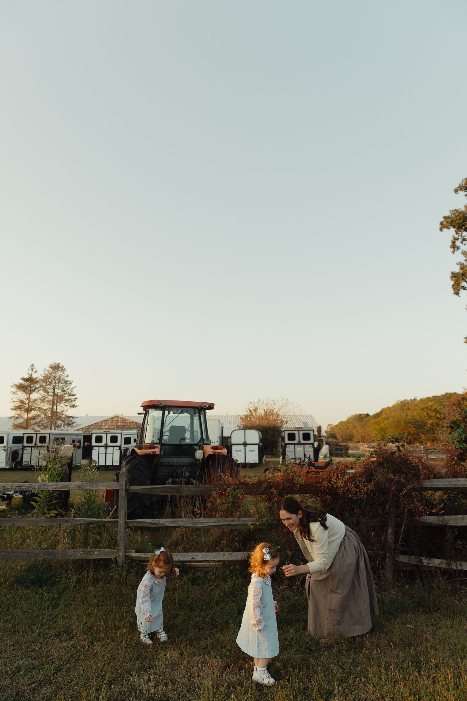 Mother tending to two young redhead daughters near wooden fence with tractor at farm.