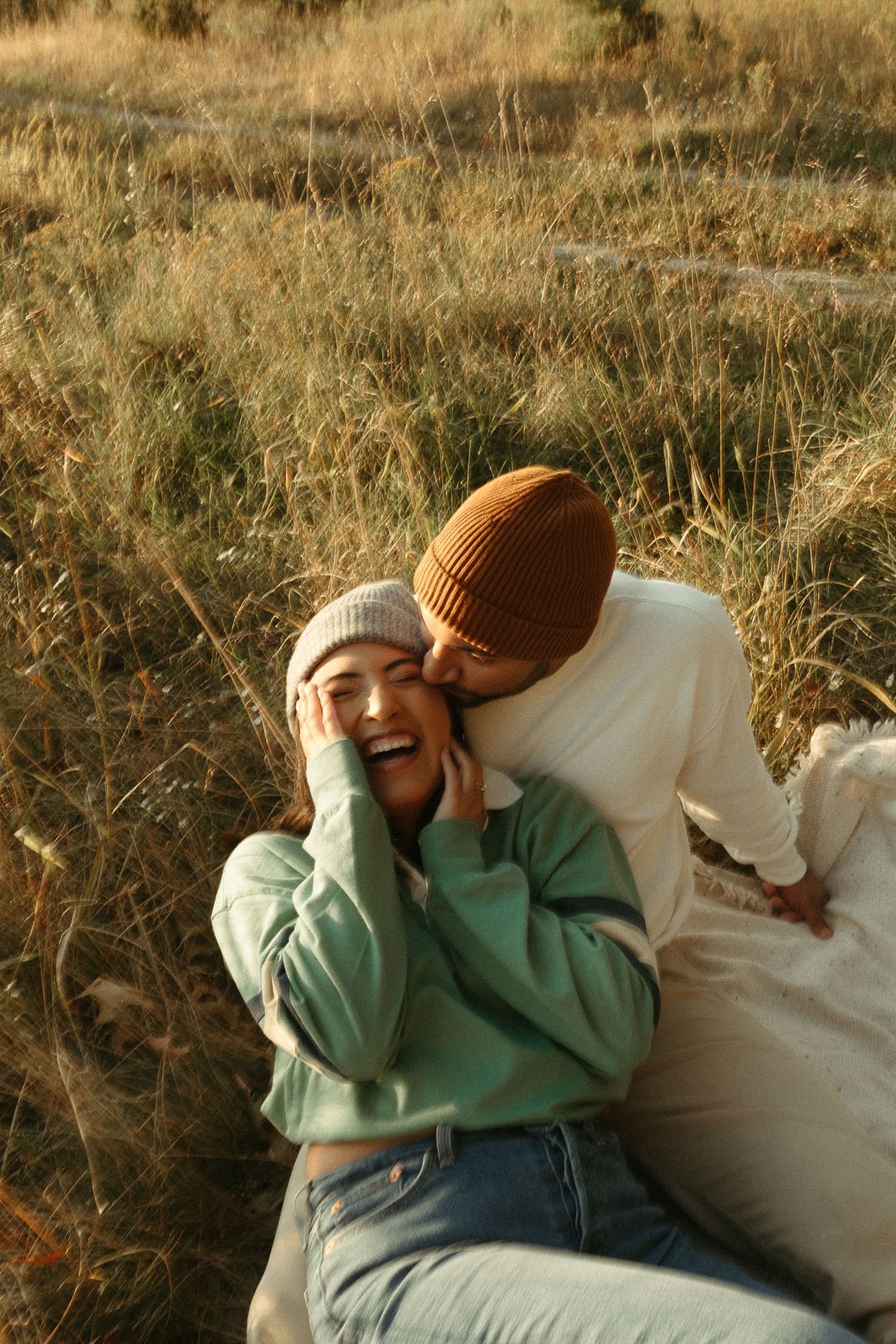 Joyful couple in beanies sharing a playful kiss in a golden autumn grass field at sunset.