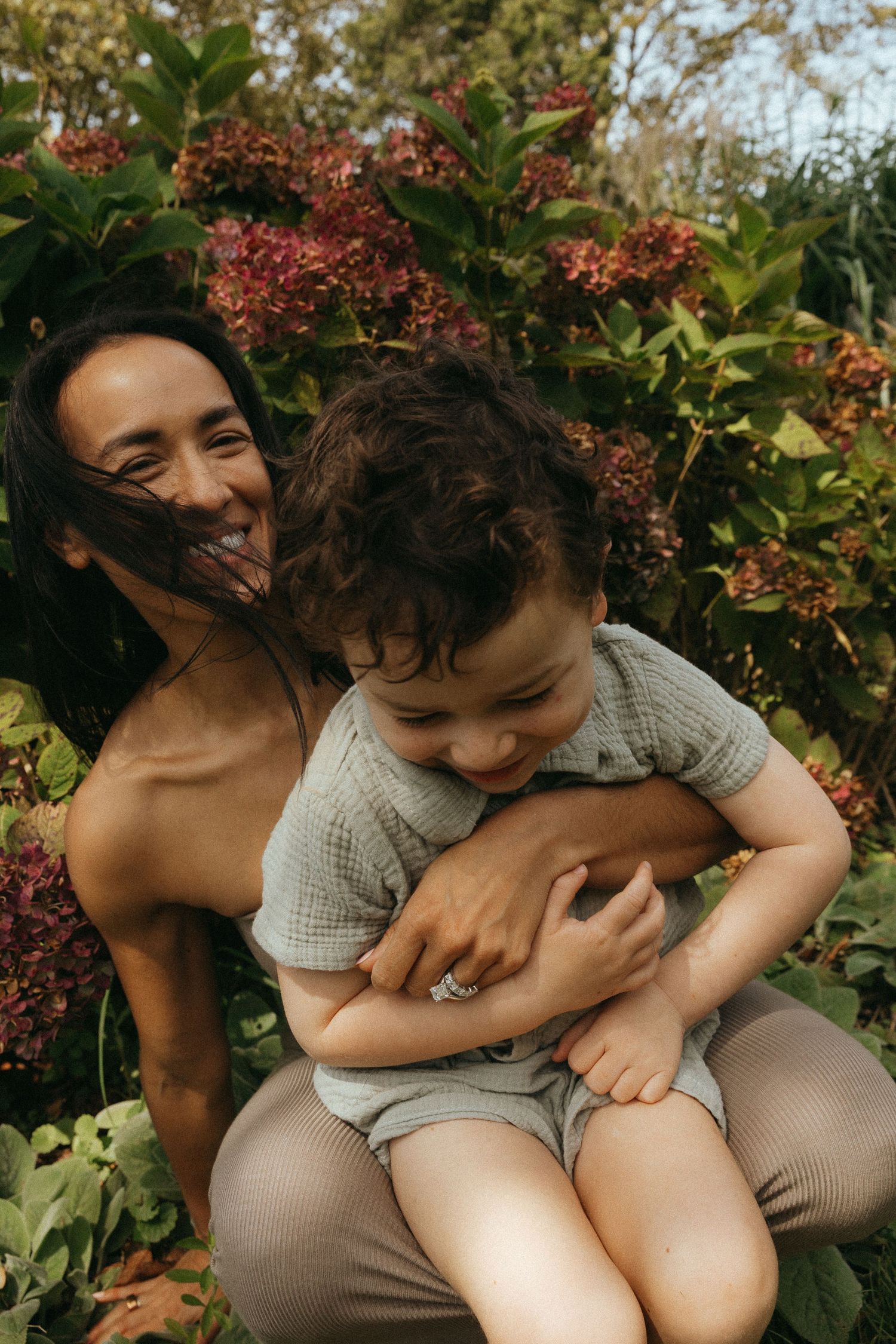Smiling mother hugs laughing toddler outdoors among lush green and burgundy garden foliage.