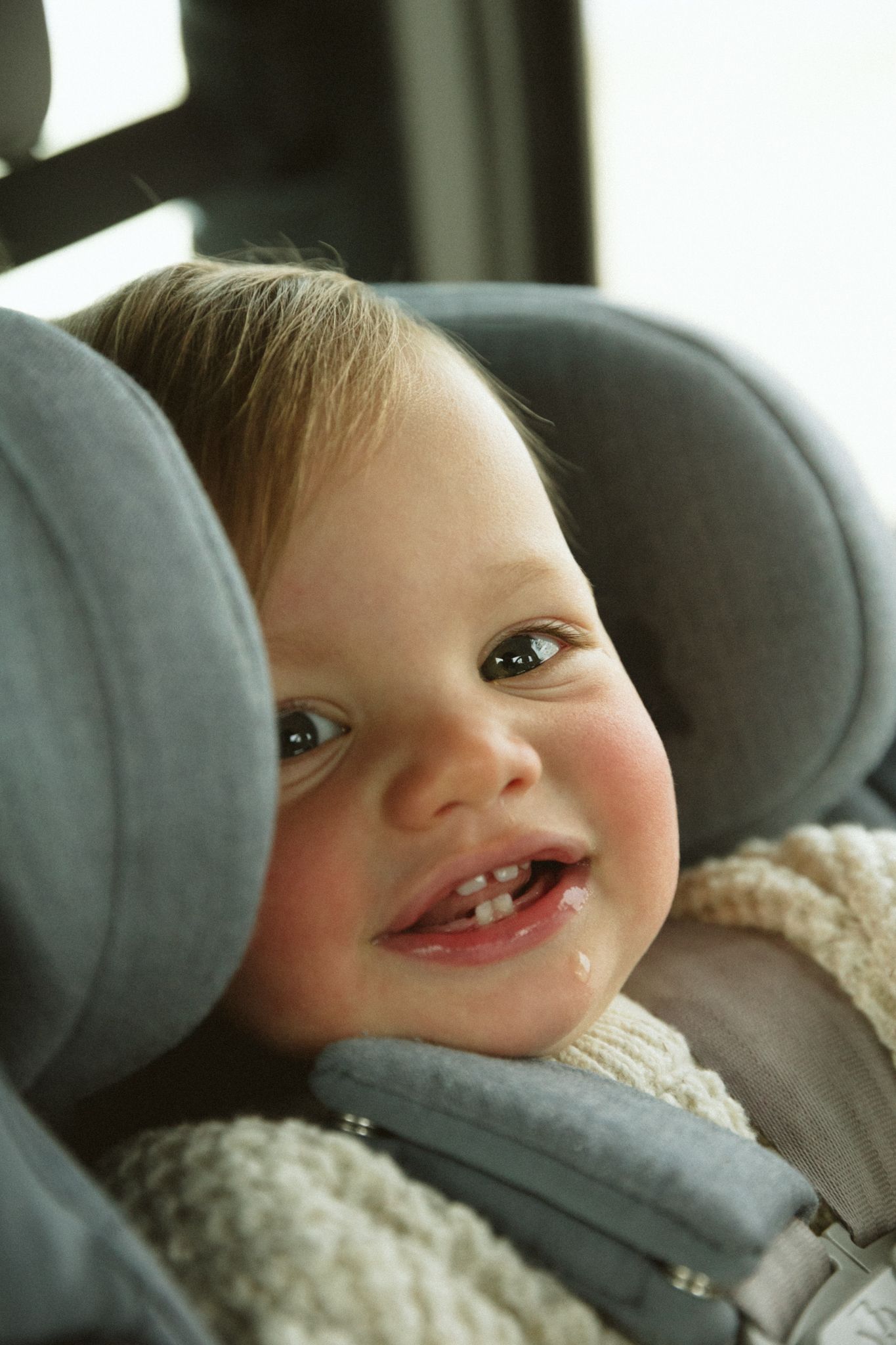 A child smiling while sitting in a grey car seat wearing a cream knit sweater.