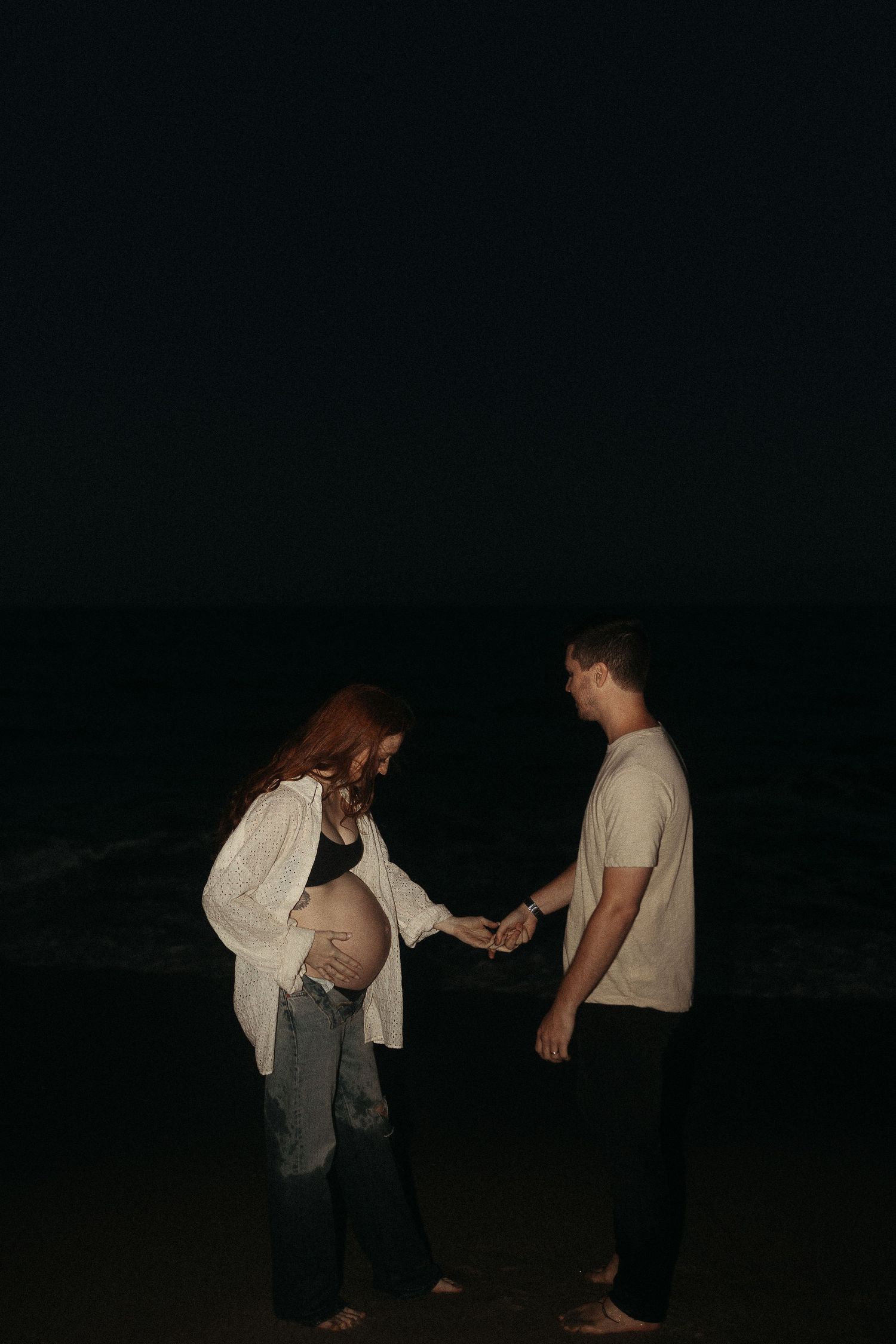 A couple shares a tender moment on a dark beach, with one wearing a flowing white cardigan.