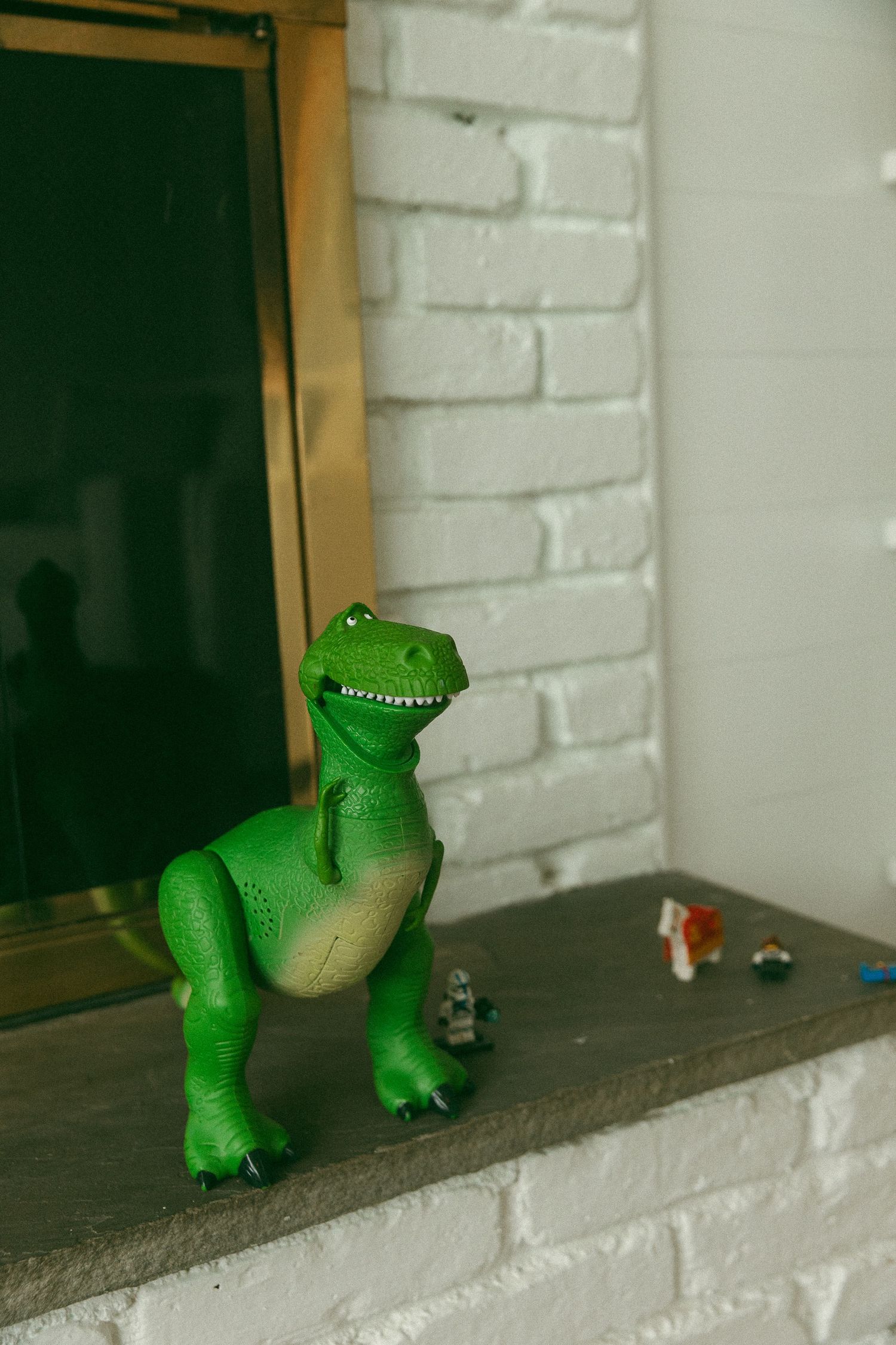 Green Rex dinosaur toy from Toy Story displayed on a fireplace hearth next to small figurines against a white brick wall.