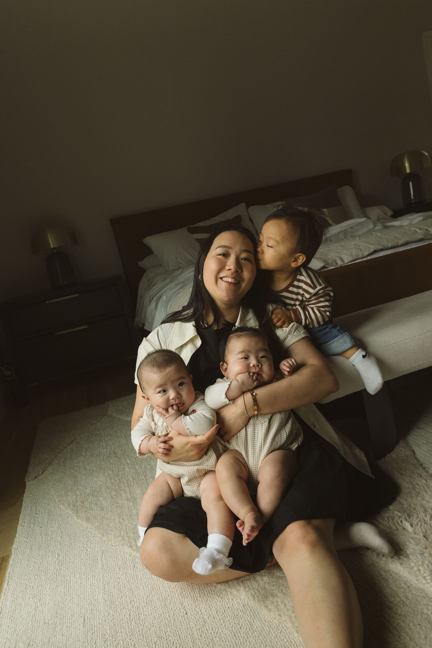 A smiling Asian mother sits on bedroom floor holding two babies while an older toddler gives her a kiss on the cheek.