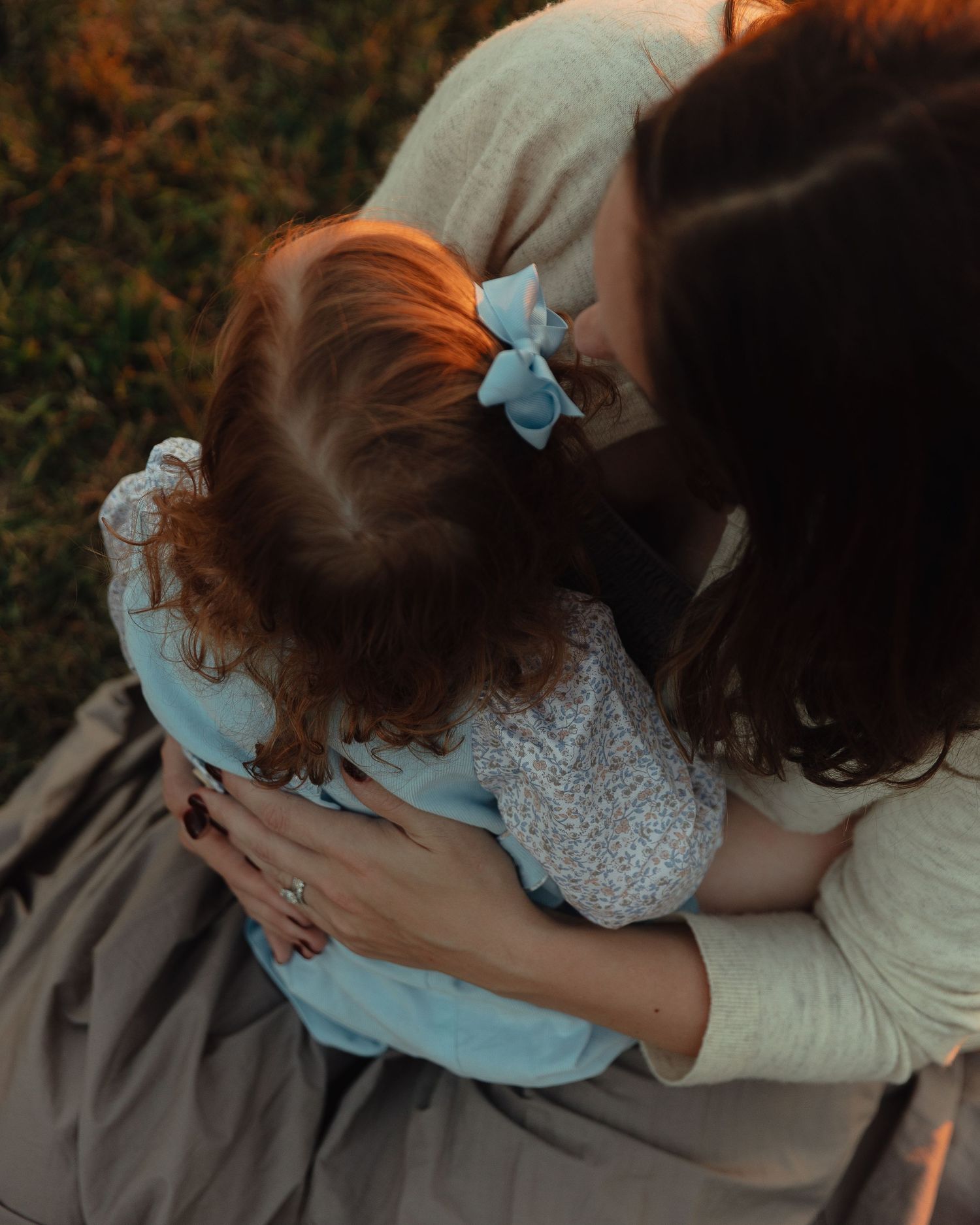 A mother tenderly embraces her young redheaded daughter in a warm outdoor setting at golden hour.