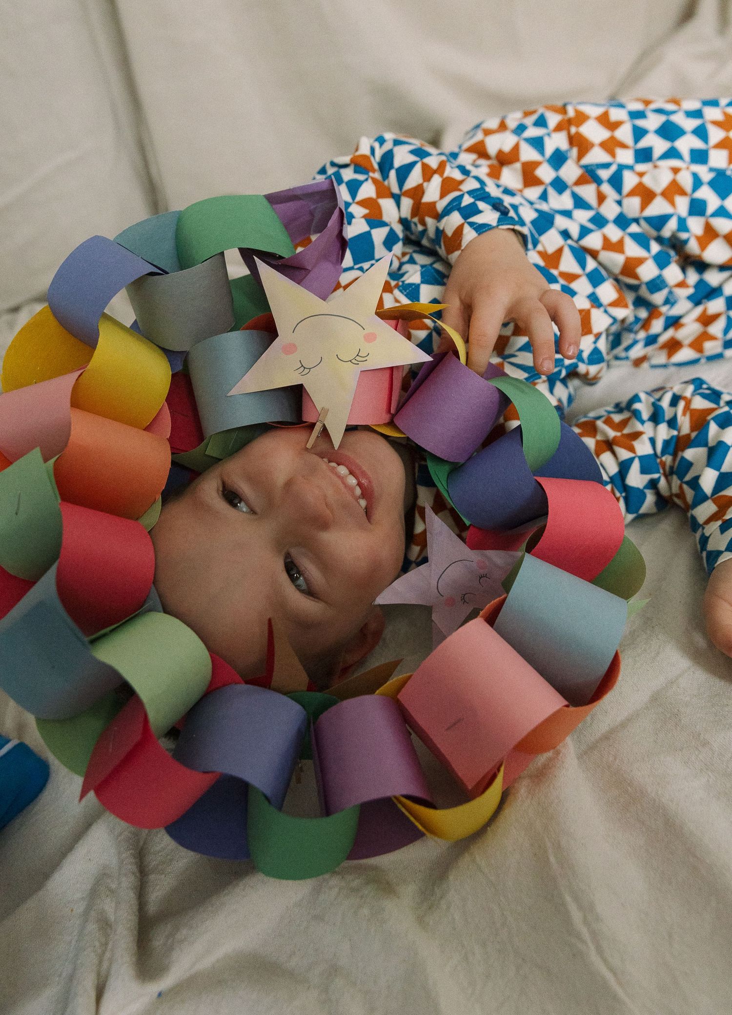 Child smiling through a colorful paper chain wreath crown, wearing patterned pajamas, holding a star cutout.
