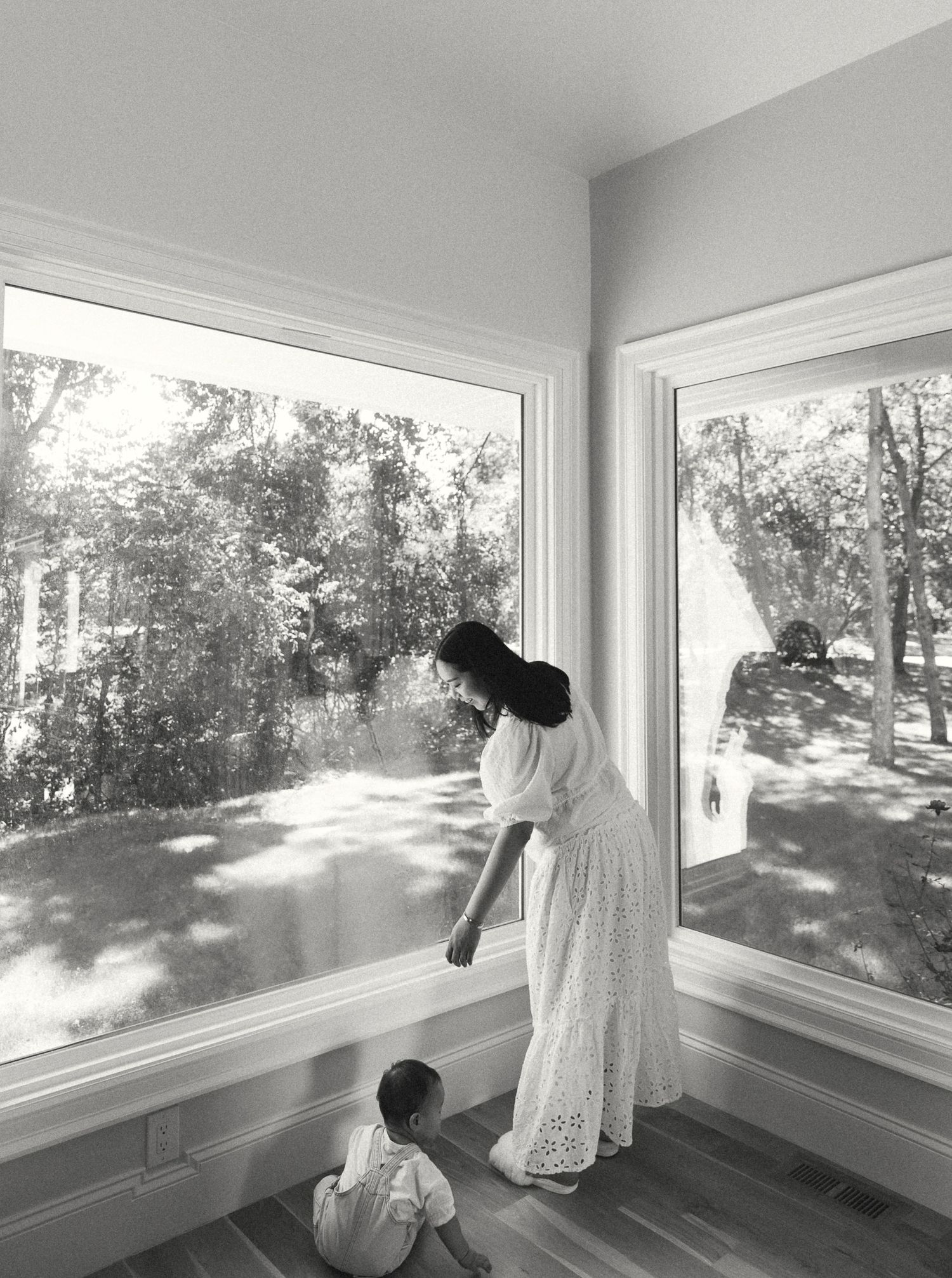 Black and white photo of a mother in a white dress leaning toward her toddler by large sunlit windows overlooking trees.