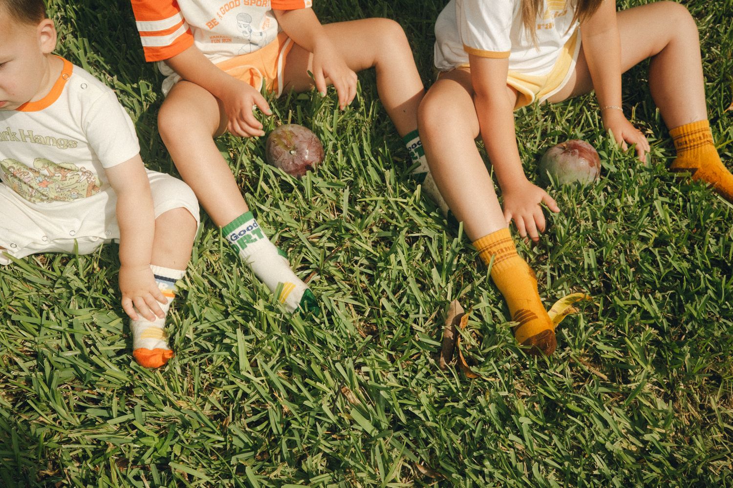 Young children sitting on green grass wearing colorful socks and casual summer outfits, playing with small balls outdoors.