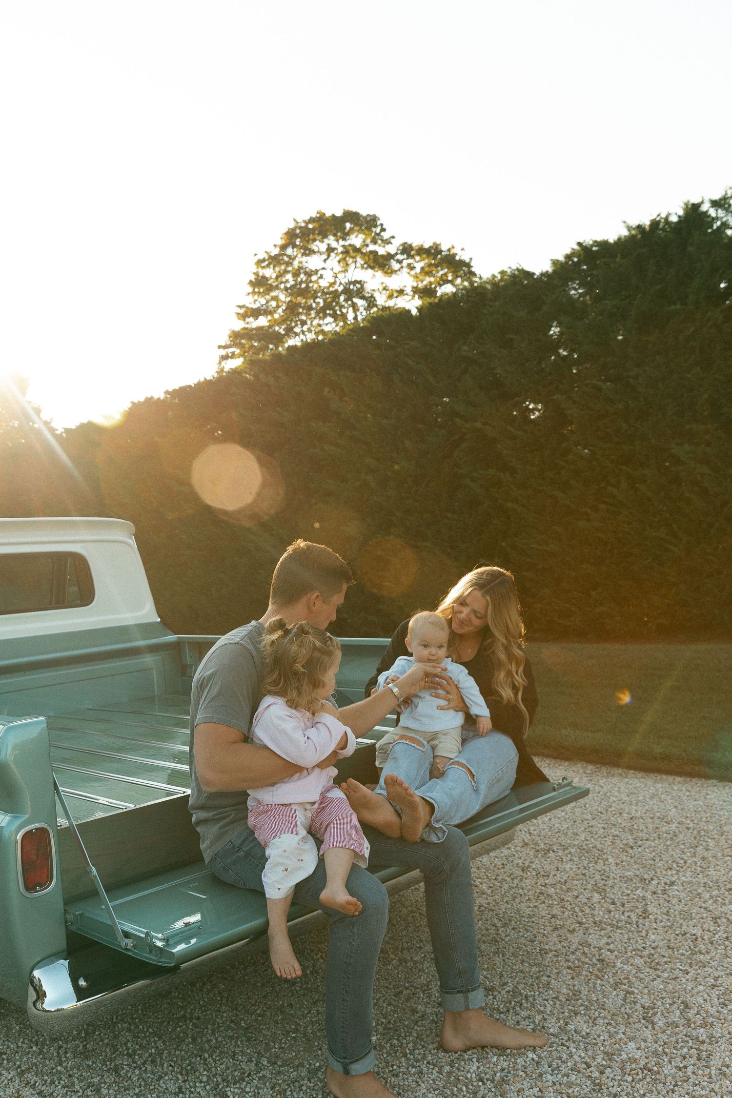 Family of four sitting on truck tailgate at sunset, parents with toddler and baby enjoying golden hour outdoors.