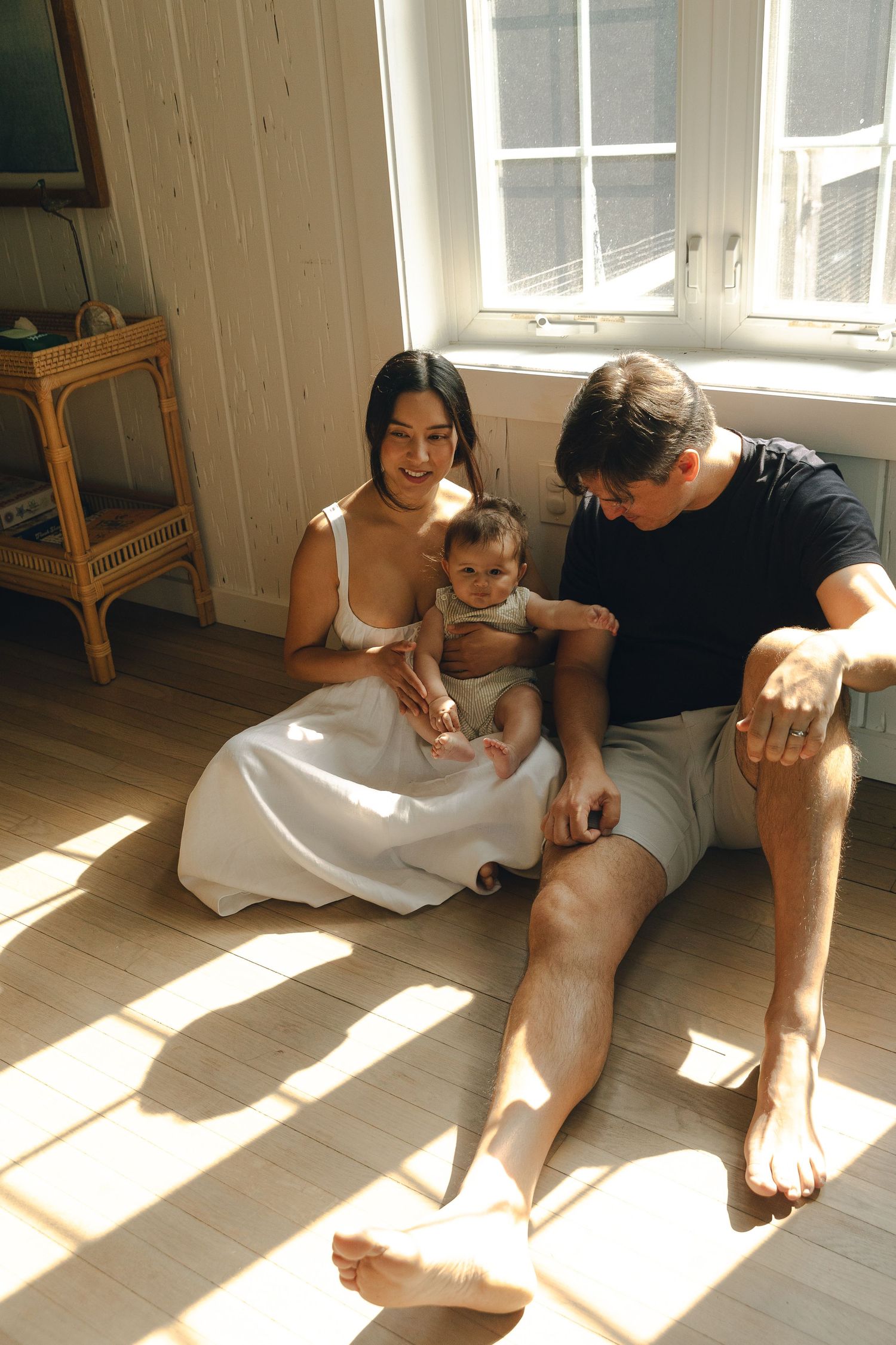 Young family sitting on sunny hardwood floor, mother in white dress holding baby, father in black t-shirt beside them.
