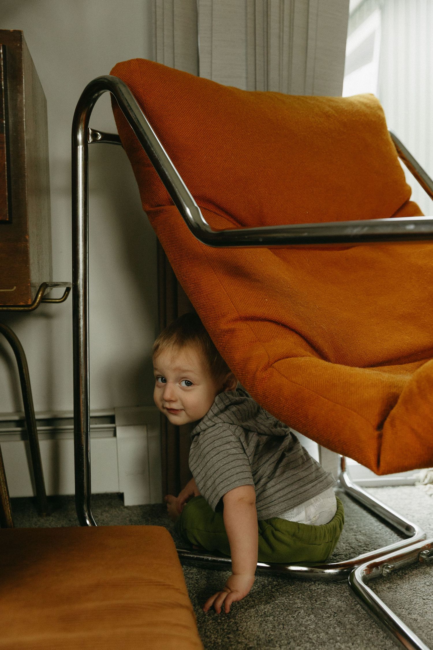 Curious baby peeking out from under orange retro chair with chrome frame indoors.