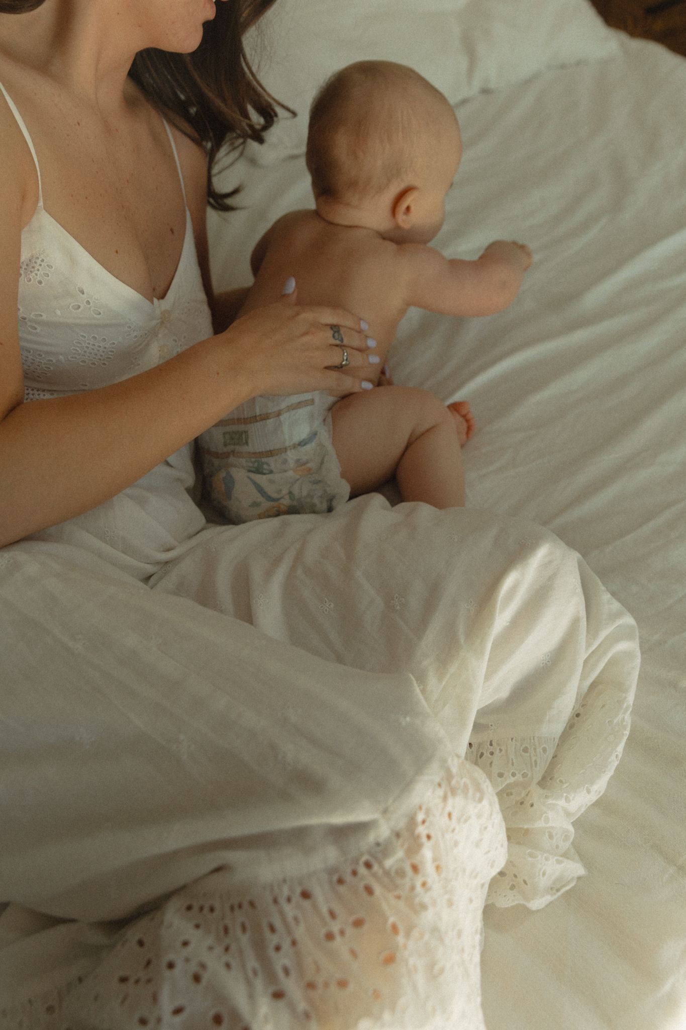 Mother in white dress sitting on bed holding baby in diaper, tender parenting moment.