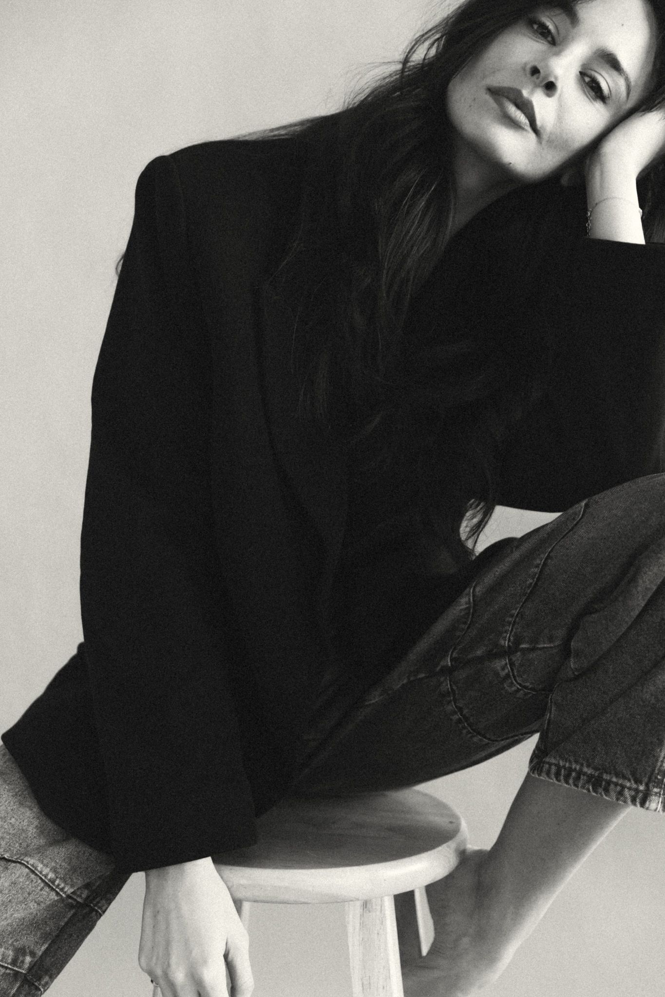 Woman in black blazer and jeans sitting on a stool, resting her head on her hand, in a black and white photo.