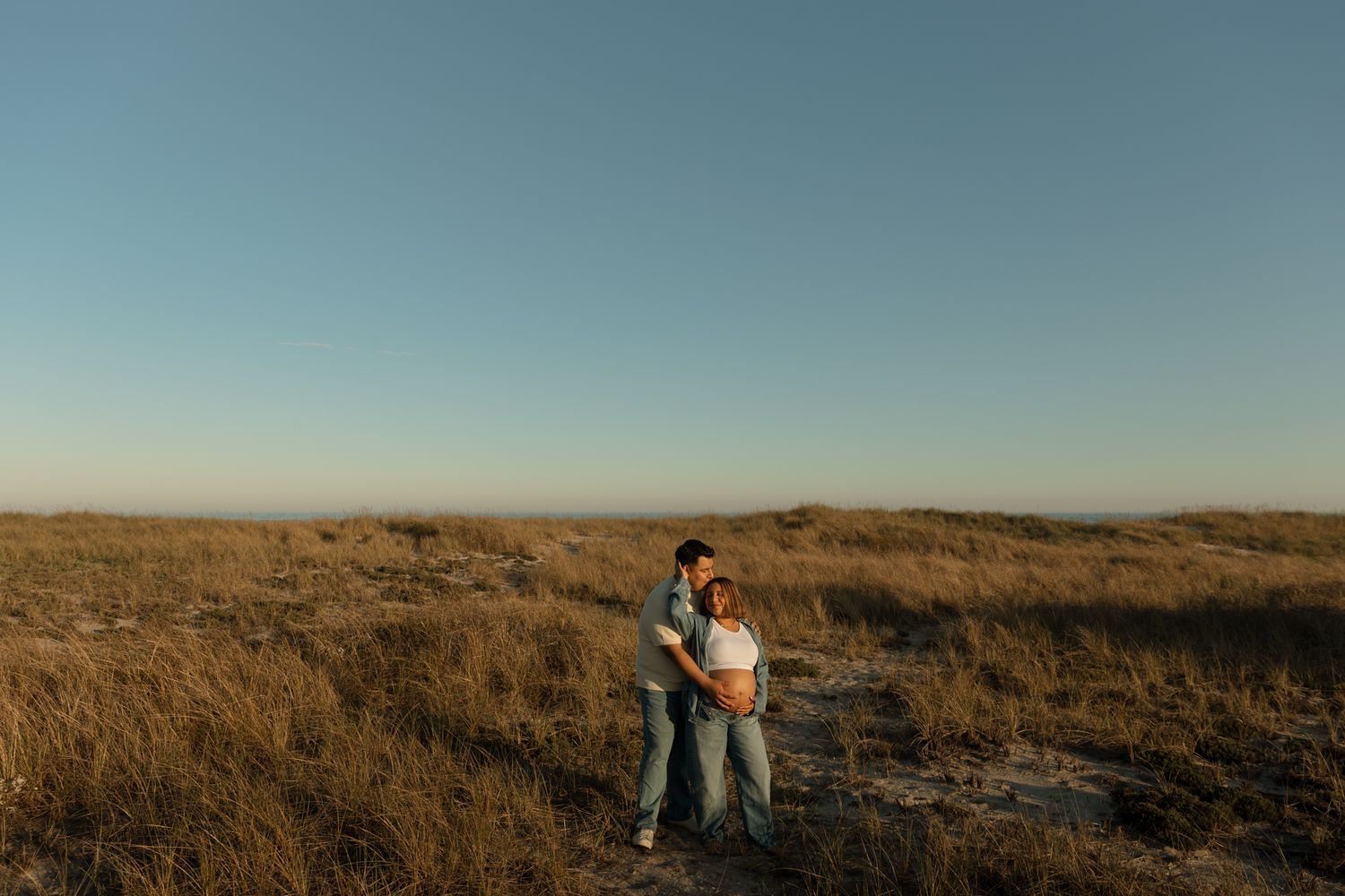 Couple embracing in golden field at sunset with clear blue sky in background.