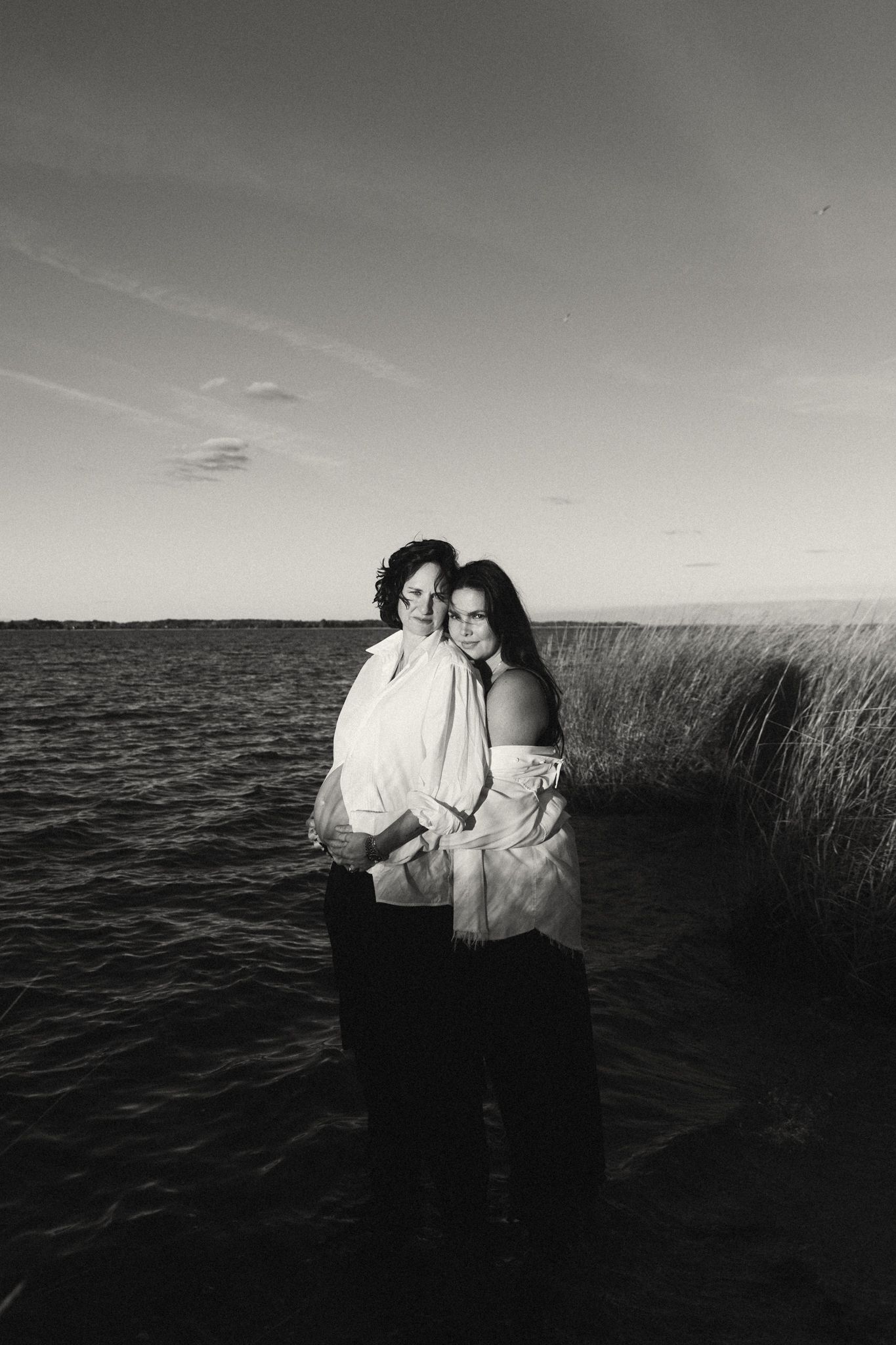 Black and white photo of two women embracing while standing in calm coastal waters at dusk.