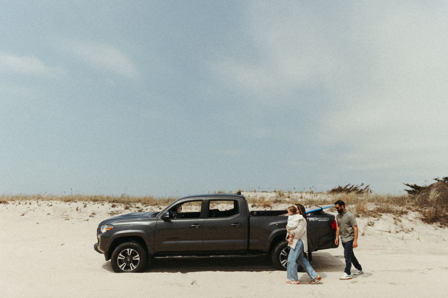 Wide angle view of a black car parked near beach dunes with people nearby.
