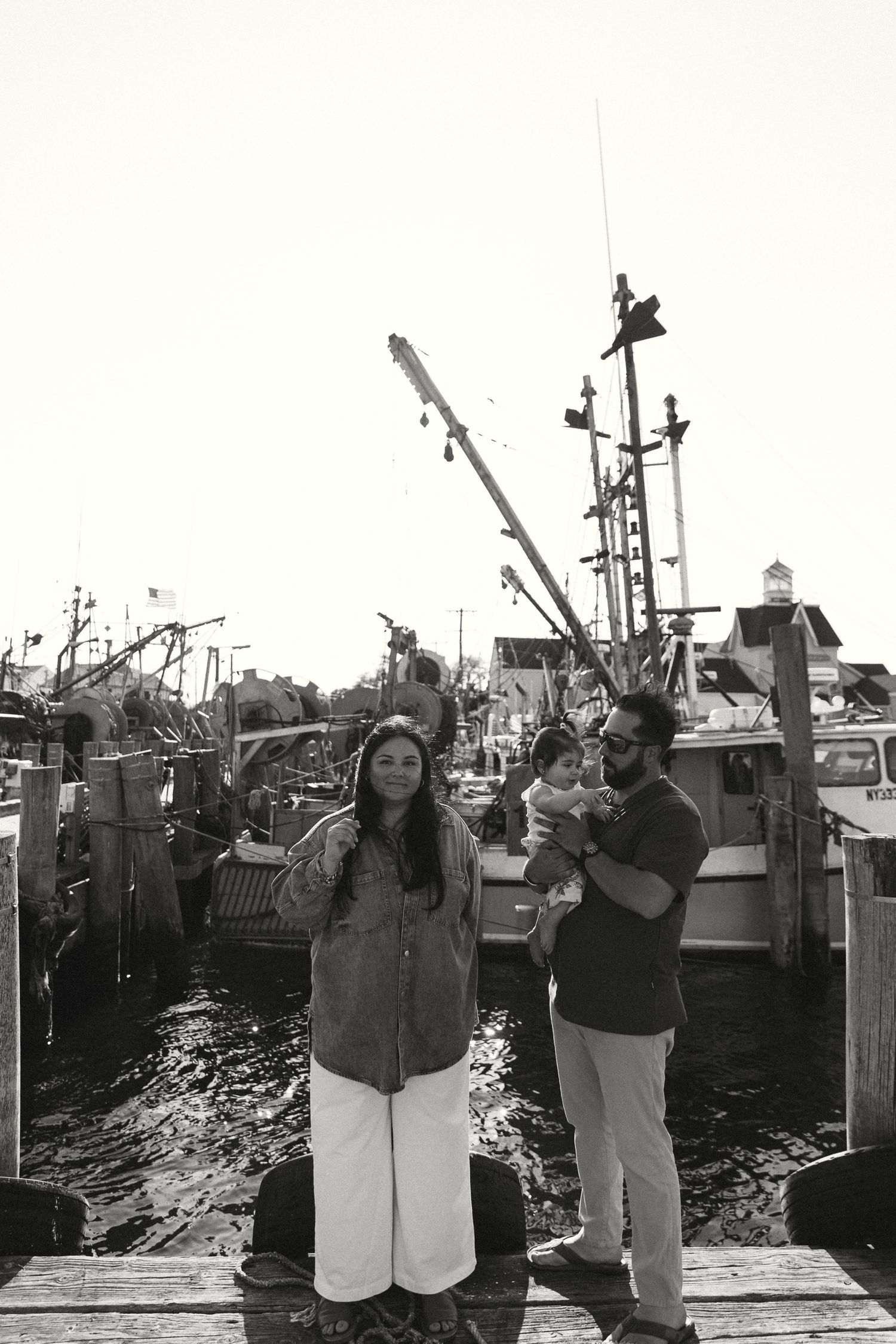 Black and white photo of people standing on a dock with fishing boats in the background.