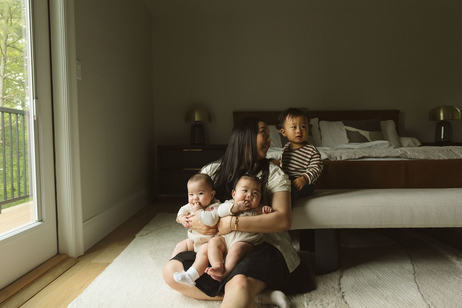 A mother sits on a bedroom floor holding two babies while an older child sits on the bed behind her.
