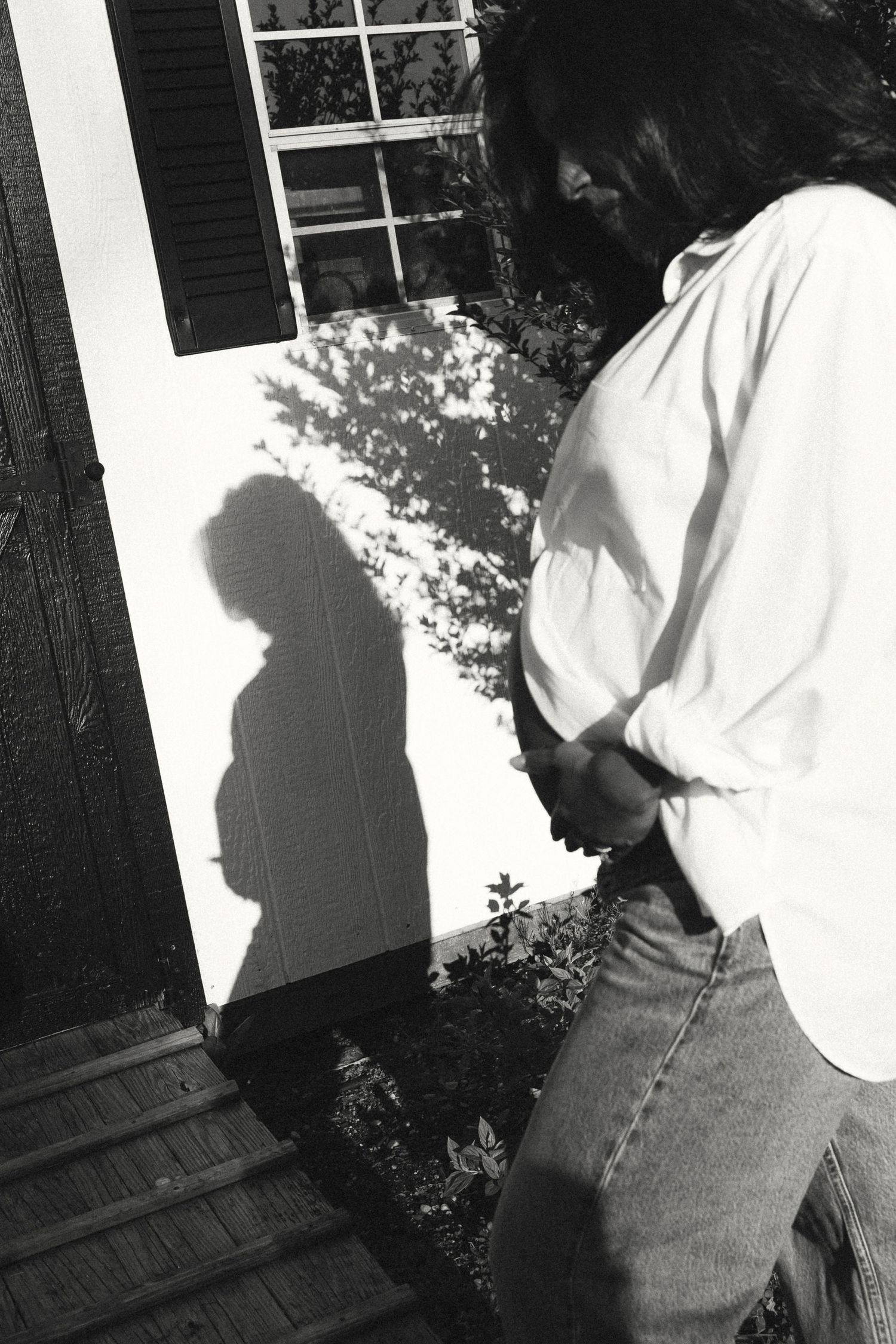 Black and white photo of a pregnant woman casting a shadow on a white wall beside a window with shutters.