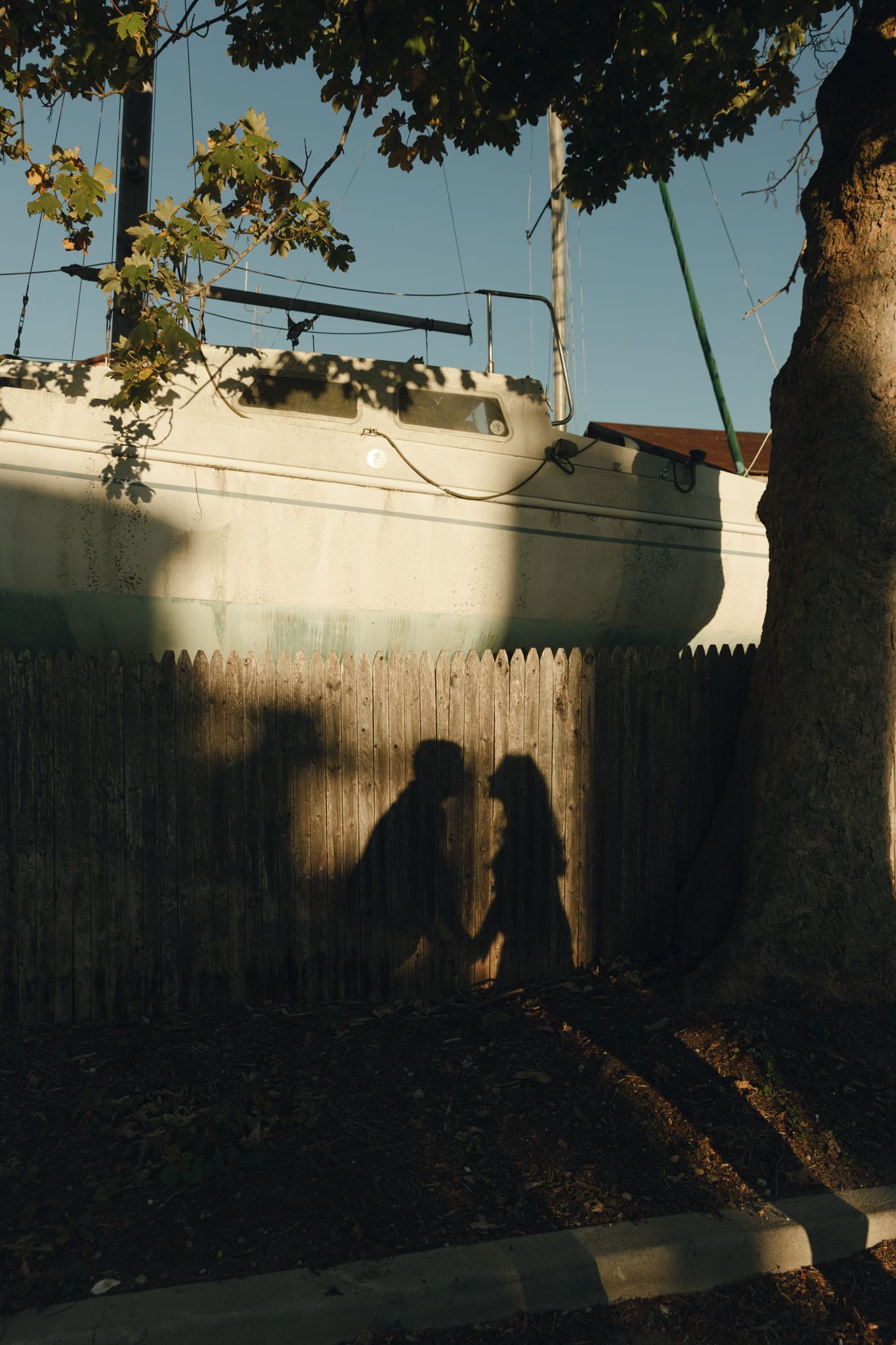 Shadows of two people holding hands cast on a wooden fence beside a sailboat in warm golden sunlight.