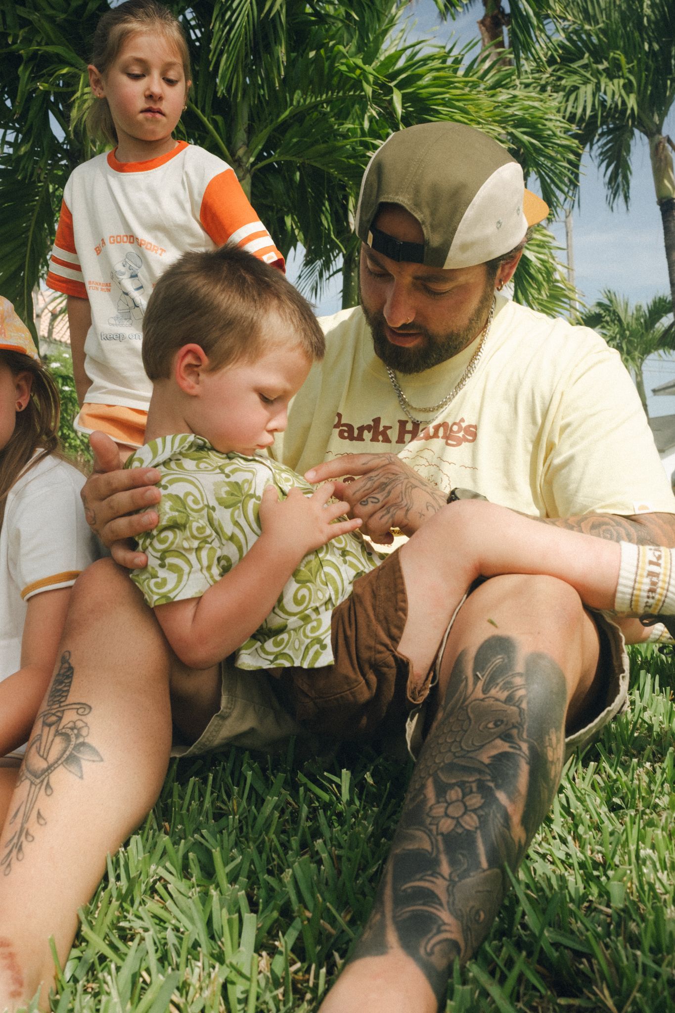 Tattooed man sitting on grass with young children near palm trees outdoors.