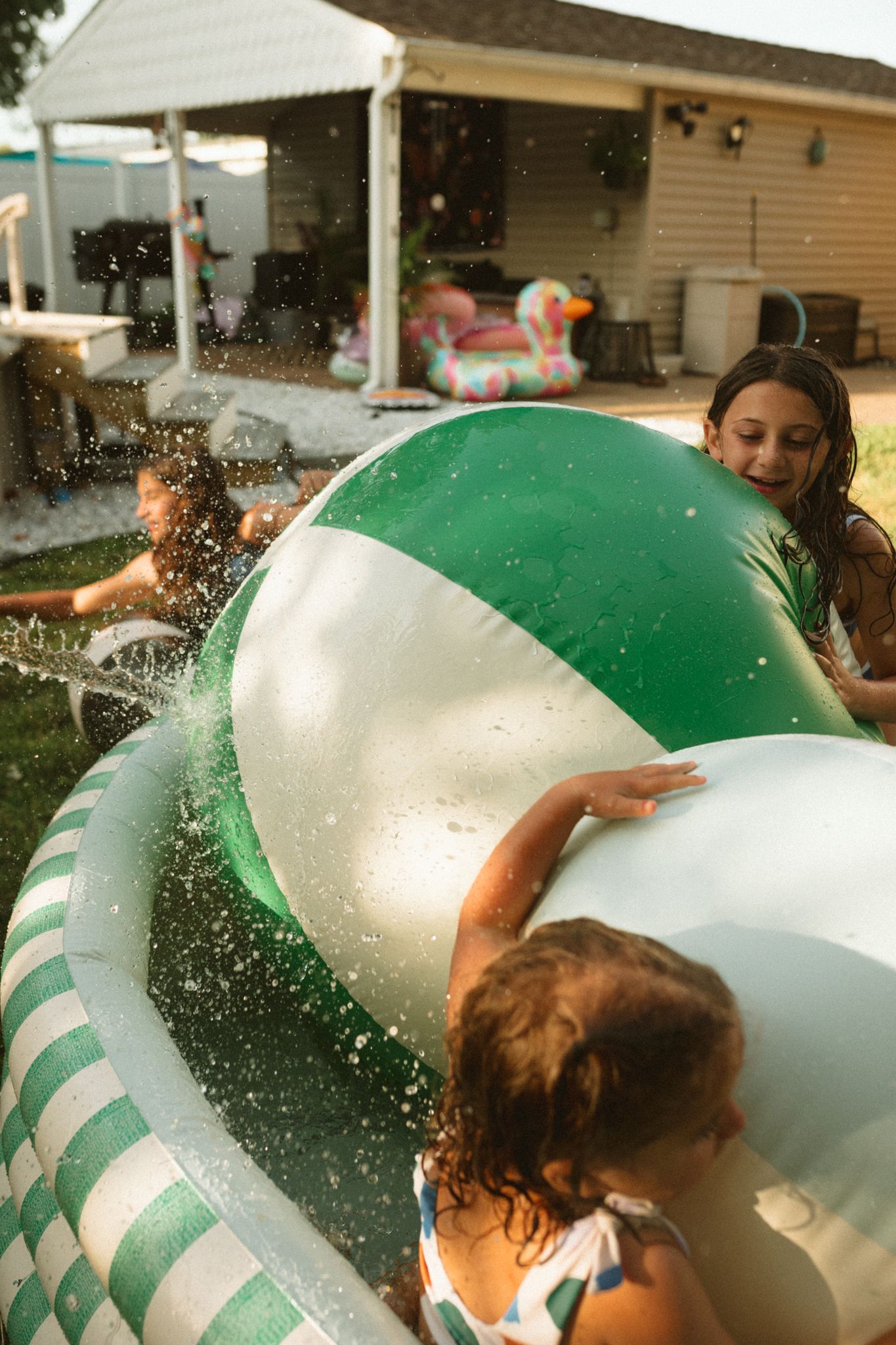 Children playing with a large green and white beach ball in a backyard splash pool.