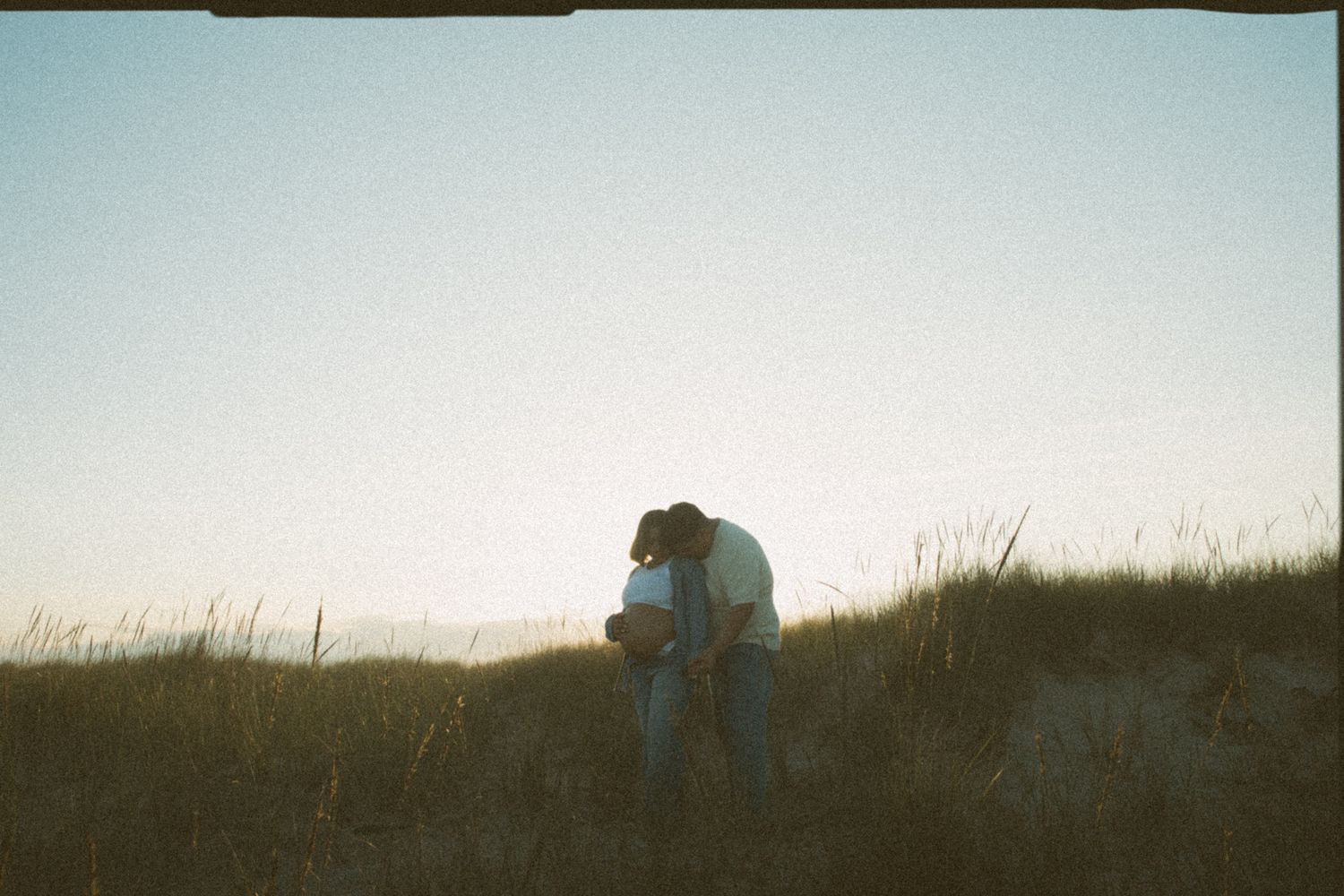 Couple embracing on sandy grass dune at twilight in romantic film photography style.