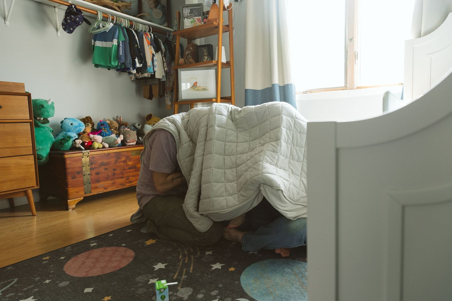 Child hiding under a quilted blanket on a colorful rug in a cozy kids bedroom with toys and clothing rack.