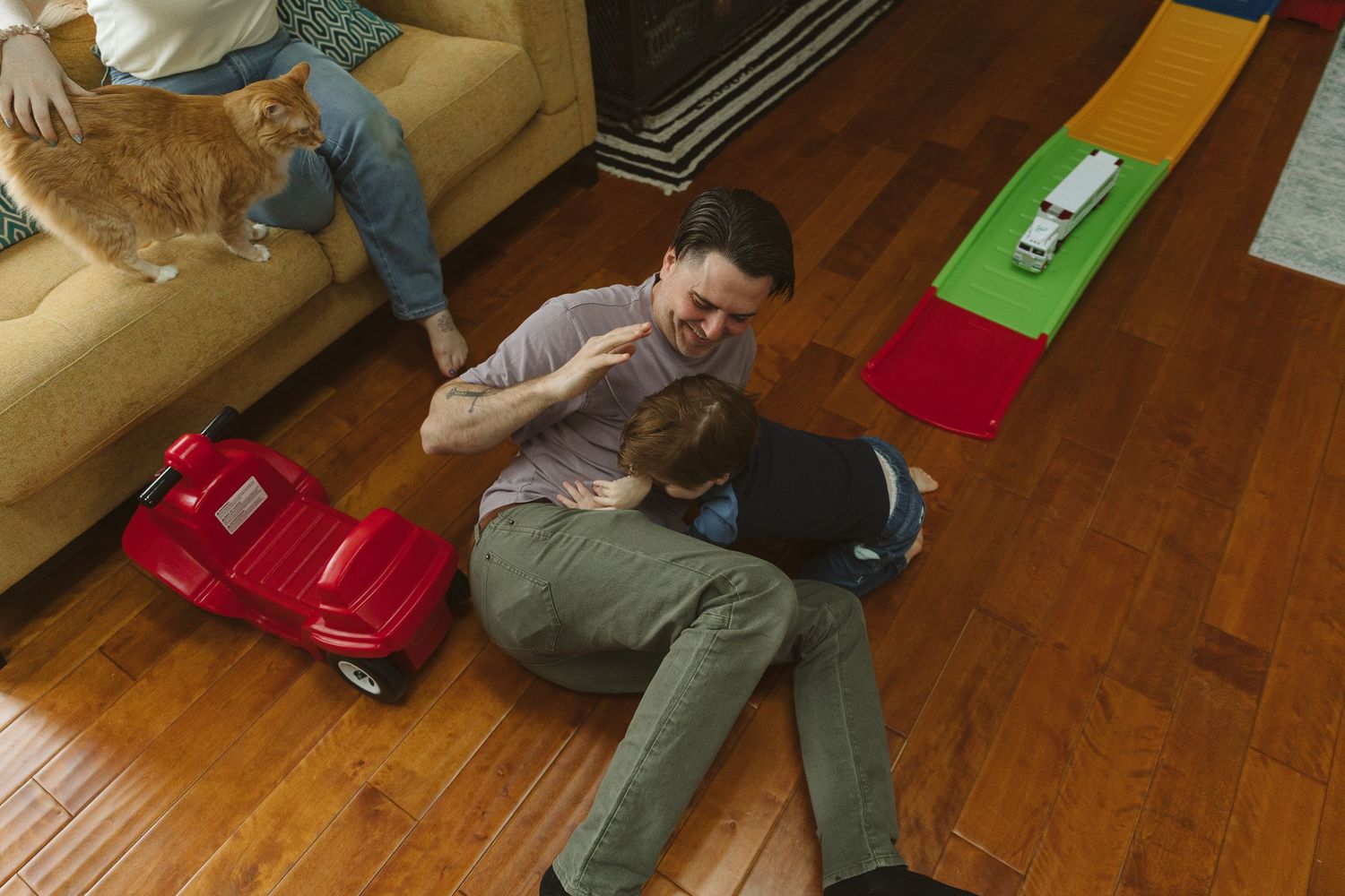 Man sitting on hardwood floor playing with child near red toy car, with orange cat on couch in cozy living room.