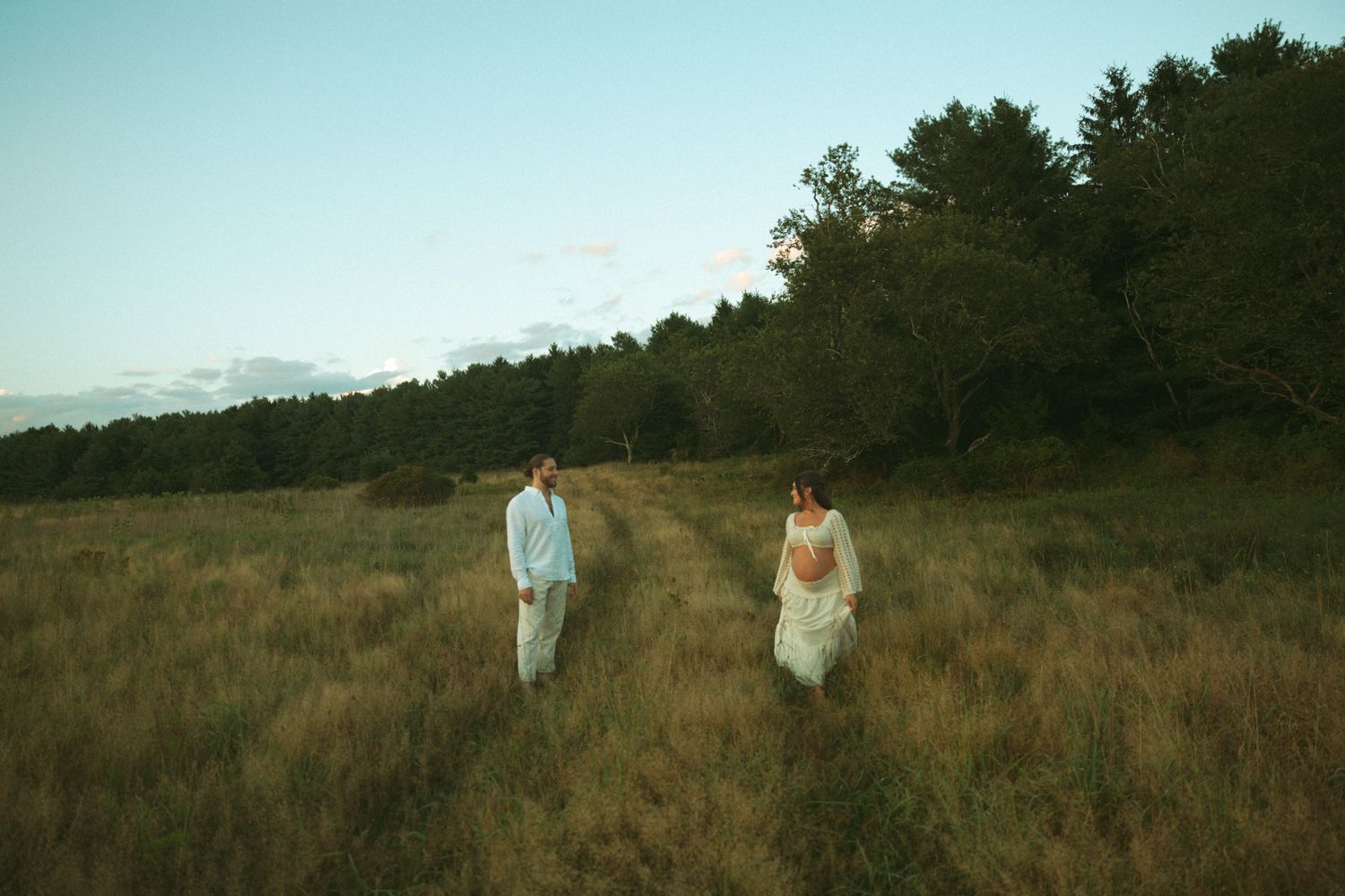 Bride and groom walk through golden meadow at dusk with lush green forest backdrop.