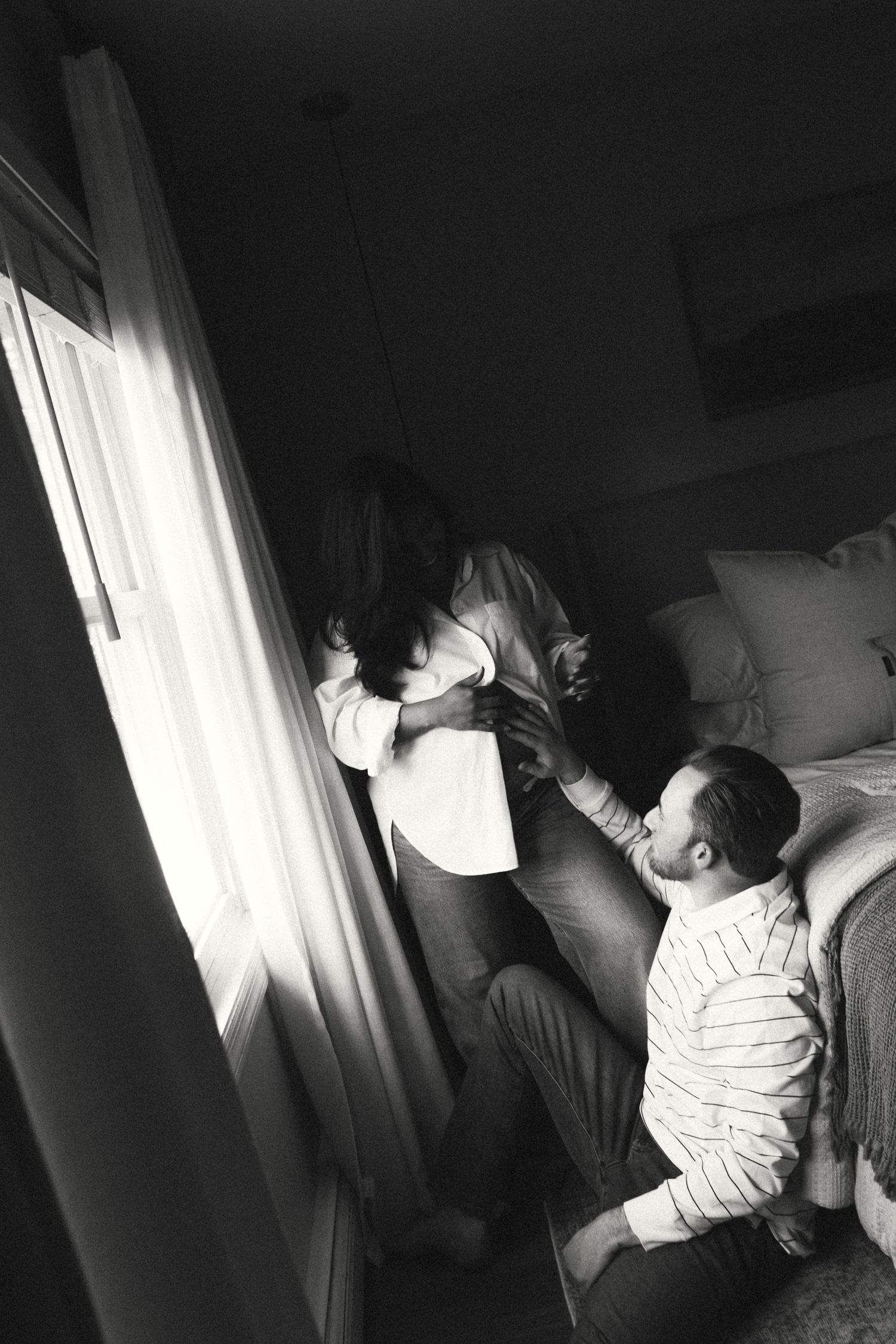 Black and white photo of a couple sharing a tender moment by a sunlit window near a bed.