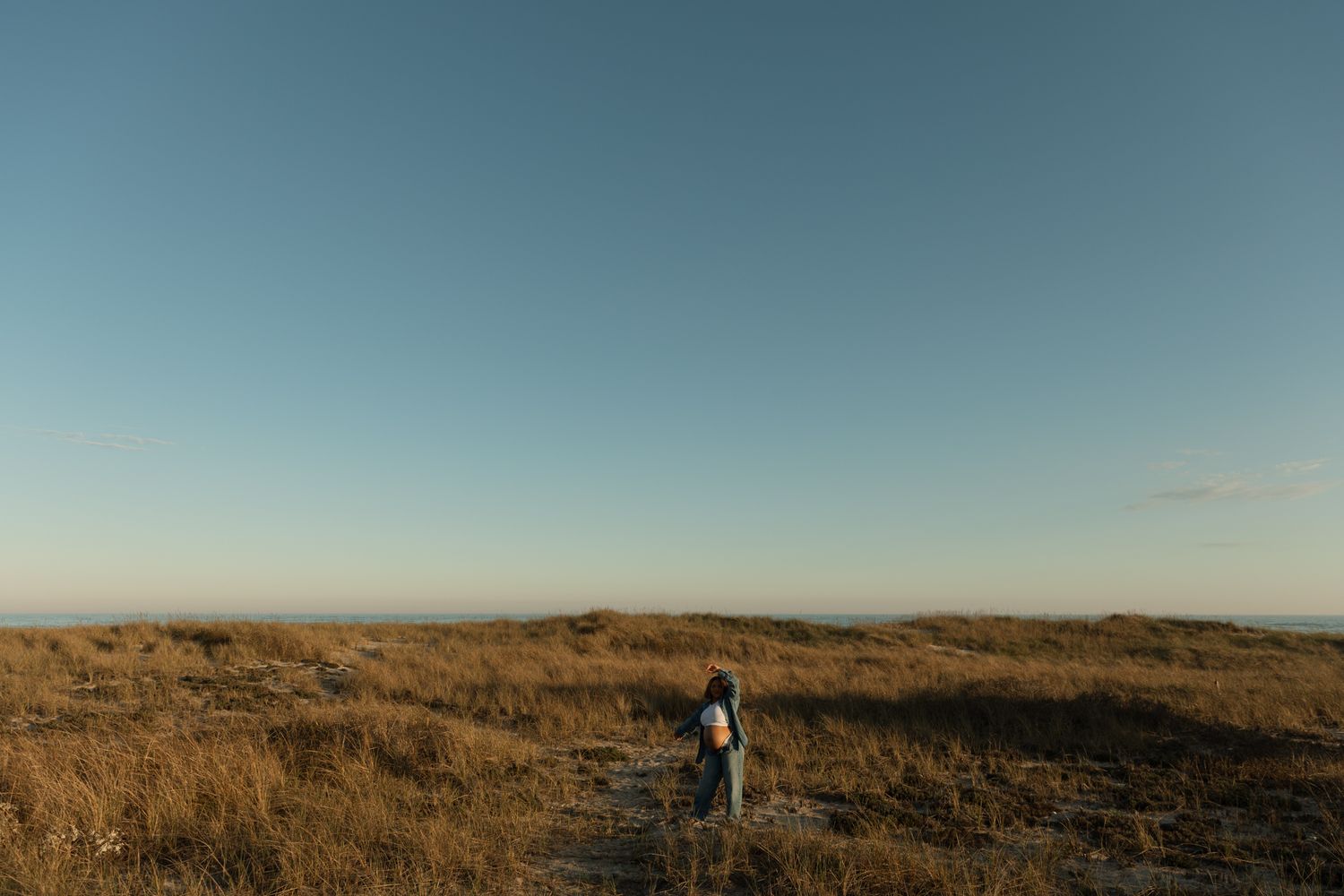 A lone hiker with a backpack walks through golden grasslands under a clear blue sky at dusk.