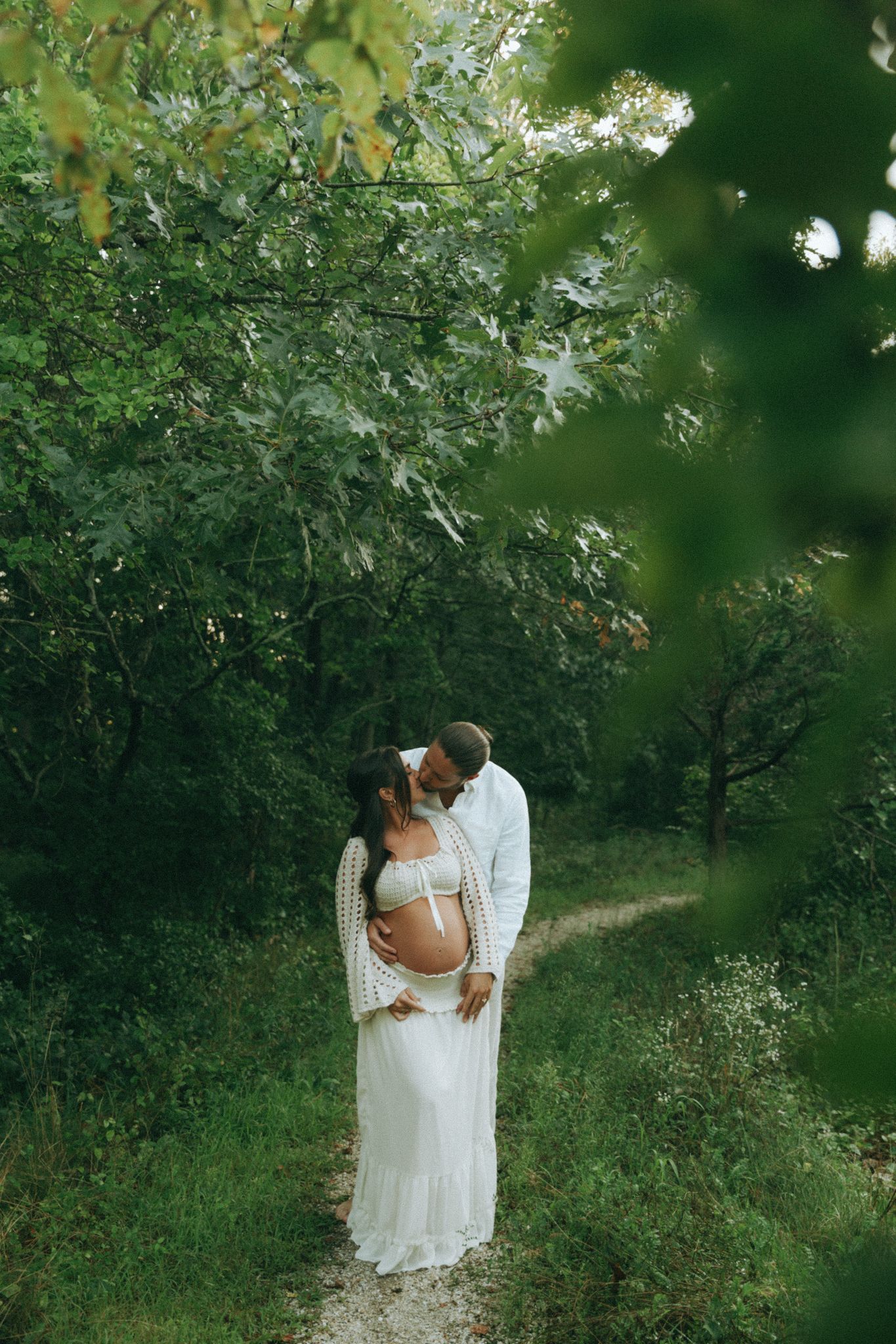 Pregnant couple embracing on a forest path, woman in white lace dress with partner in white shirt among lush green trees.