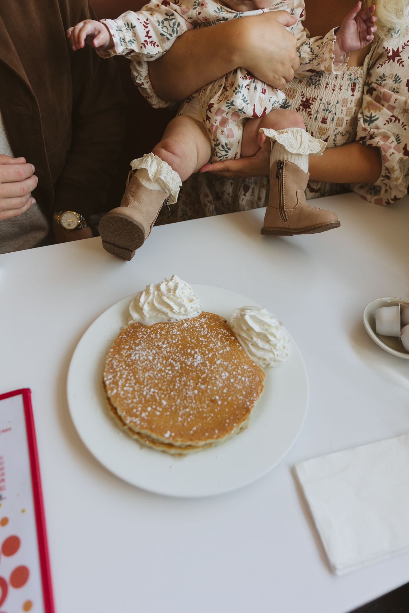 Fluffy pancakes topped with whipped cream and powdered sugar on a white plate, with two young children sitting at a restaurant table.