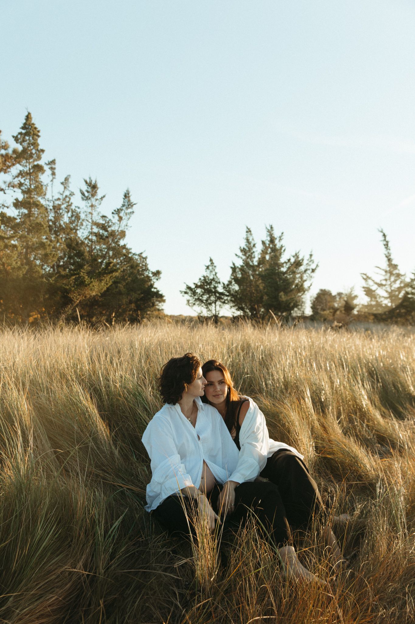 Two women sitting closely together in golden grass field with pine trees at sunset.