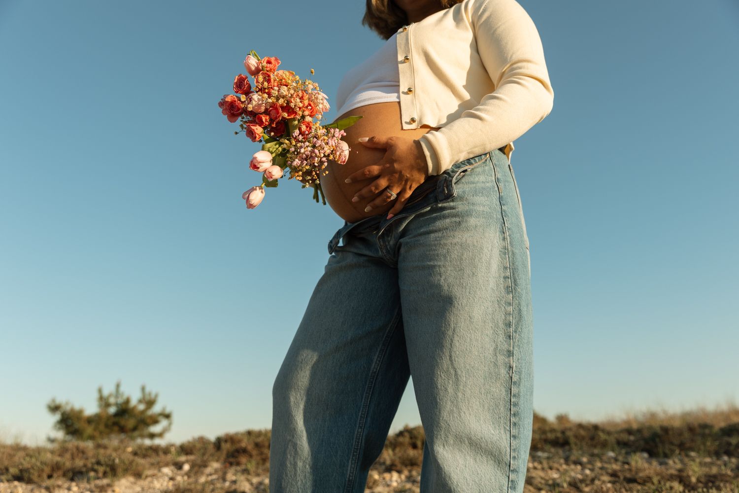 Pregnant woman in white crop top and jeans holds colorful wildflower bouquet against blue sky.