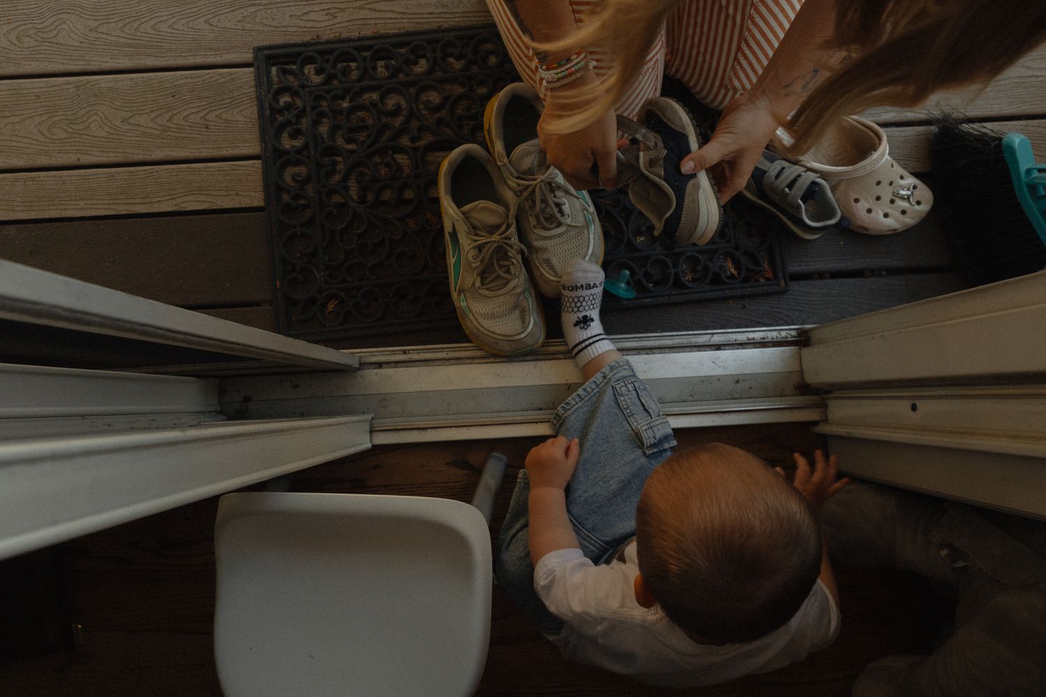 Overhead view of baby reaching toward doormat with shoes while adult holds sneakers and crocs at home entrance.