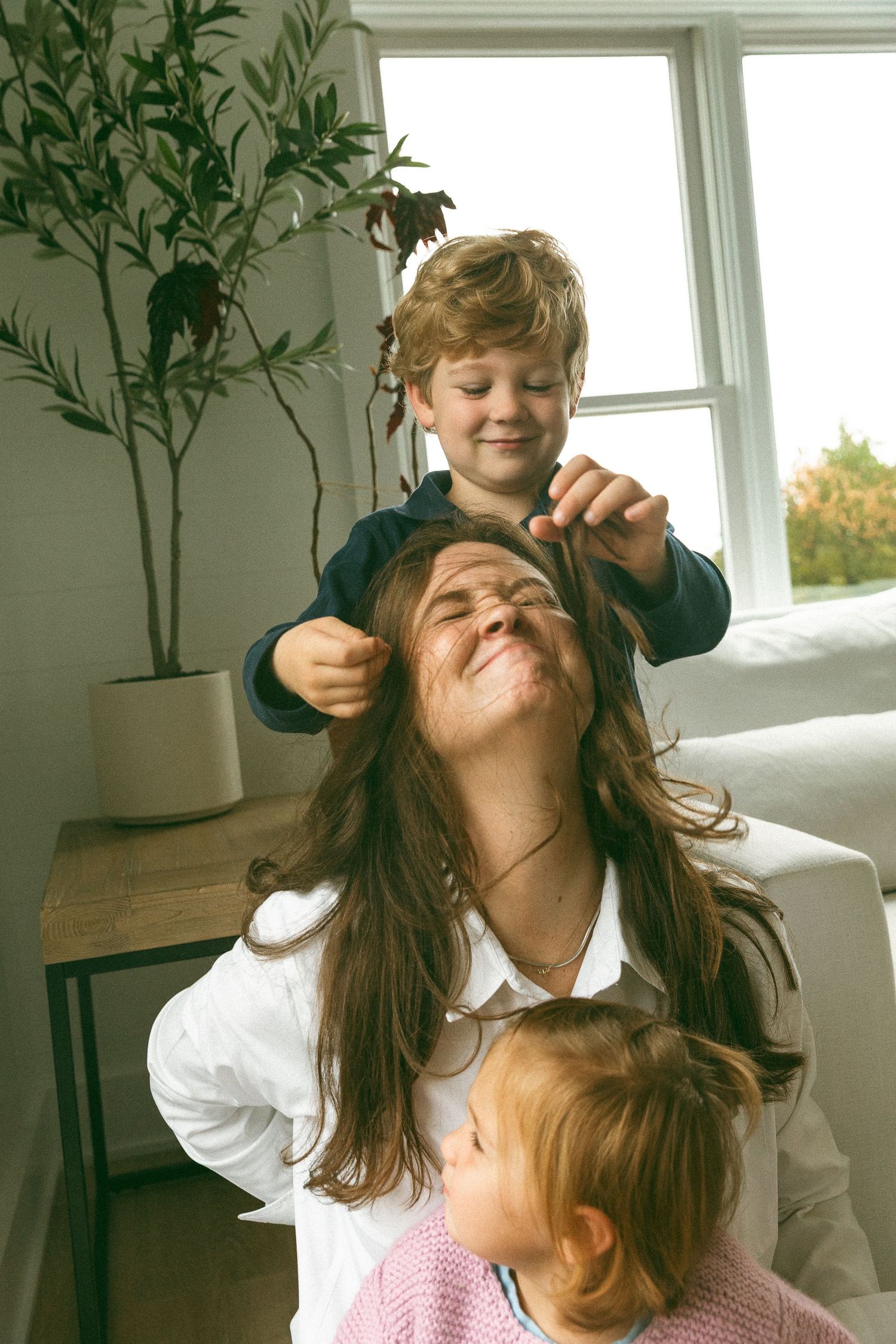 A joyful mother laughs as her young son plays with her long hair while a toddler watches nearby indoors.
