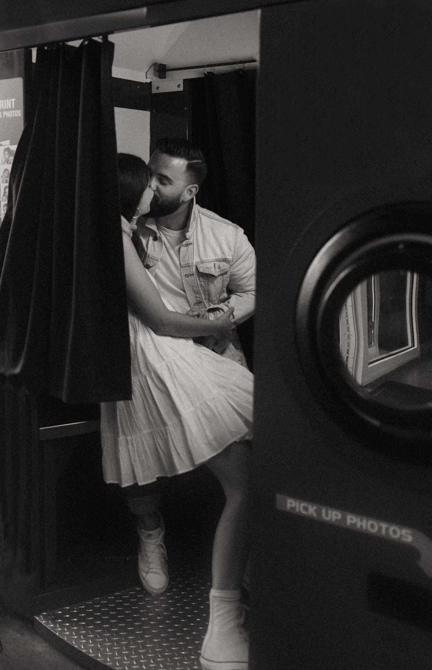 Black and white photo of a couple embracing and kissing inside a photo booth with a pick up photos sign.
