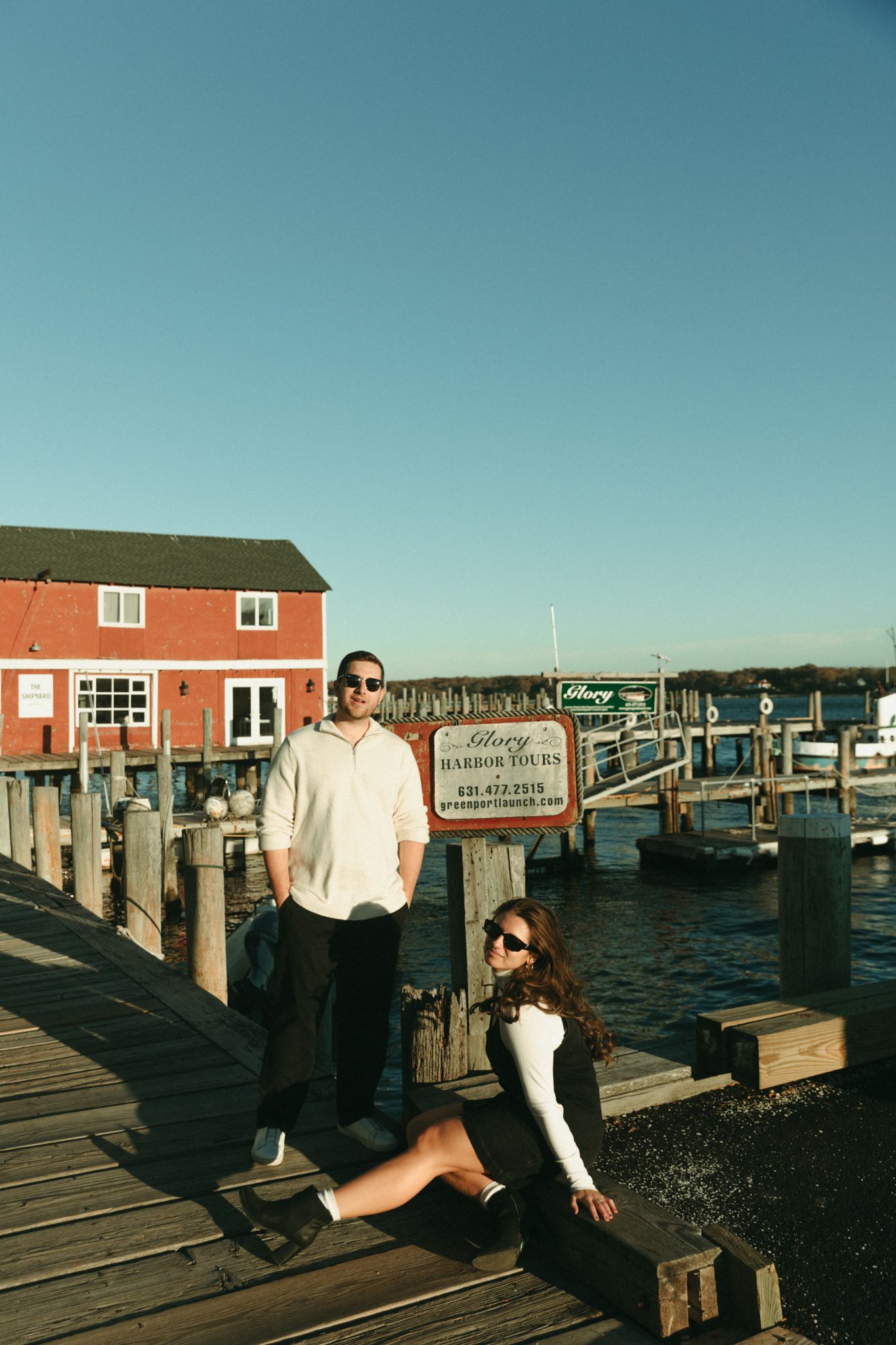Couple posing on a wooden dock near Glory Harbor Tours sign with a red building and marina in the background.