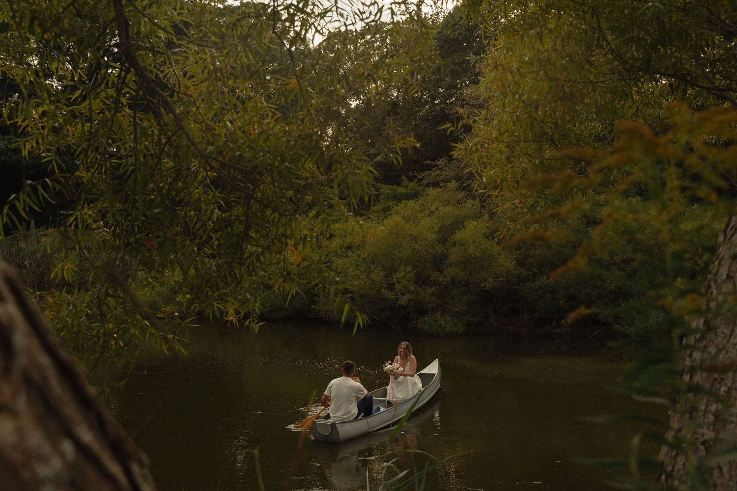 Couple rowing a small white boat on a serene tree-lined river in soft evening light.