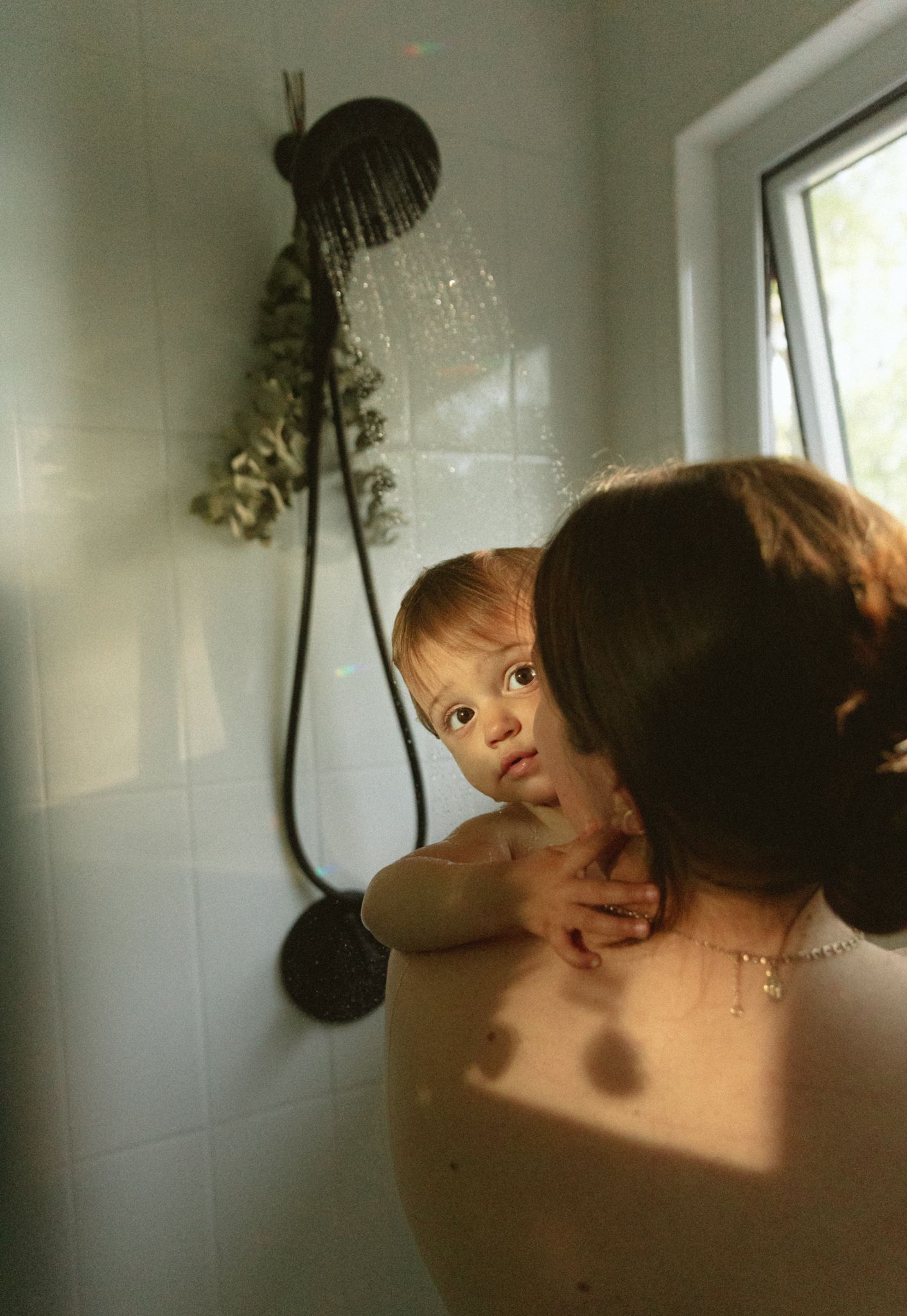 Mother holding toddler in shower with running water, white tiles, and natural light from window.